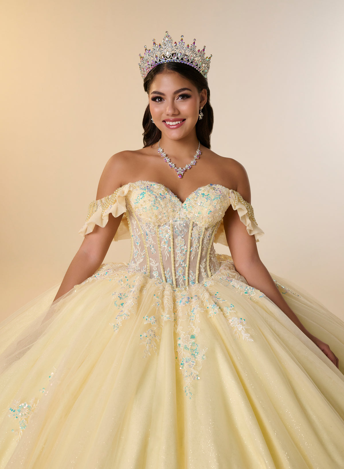 A woman wearing a sparkling off-the-shoulder ball gown in pale yellow with light blue and silver floral embroidery, a matching tiara, and a pearl necklace, posing against a warm beige studio background. / Romantic off-shoulder ball gown with delicate embellished appliqués, a fitted bodice, a voluminous tulle skirt, and a statement bow at the back.