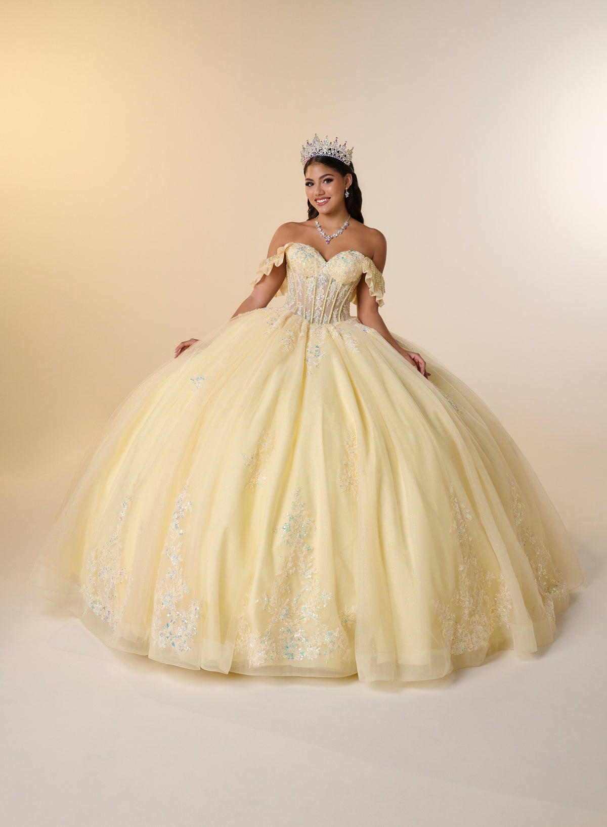 A woman wearing a voluminous pale yellow quinceañera ball gown with off-the-shoulder straps, a beaded sweetheart bodice, and embroidered tulle details, posed against a warm beige studio backdrop. / Romantic off-shoulder ball gown with delicate embellished appliqués, a fitted bodice, a voluminous tulle skirt, and a statement bow at the back.