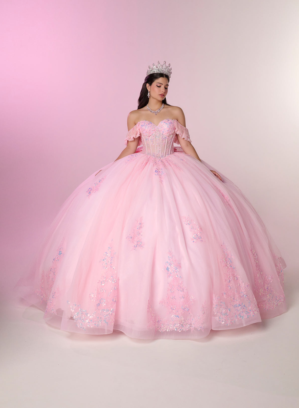 A woman wearing a voluminous light pink ball gown with off-the-shoulder puffed sleeves, a fitted embellished bodice, and a full layered tulle skirt with floral embroidery, posed against a soft pink studio backdrop. / Romantic off-shoulder ball gown with delicate embellished appliqués, a fitted bodice, a voluminous tulle skirt, and a statement bow at the back.