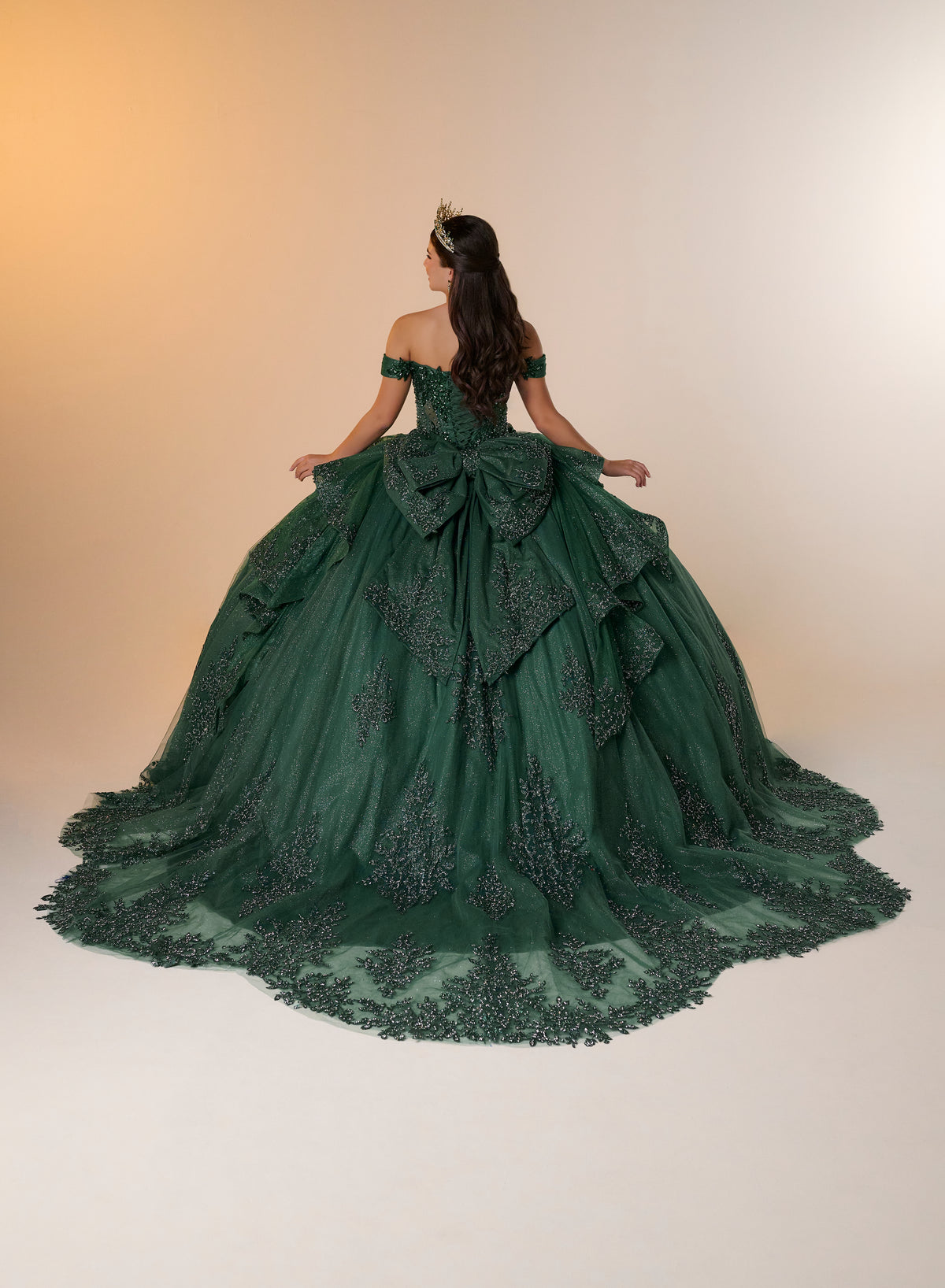 A woman stands with her back to the camera wearing an off-the-shoulder dark green ball gown with a large bow at the back and a long train, photographed against a warm beige studio background. / Luxurious off-shoulder ball gown featuring glittering embellishments, layered draped skirt details, and a dramatic cathedral-length train for a breathtaking princess-inspired look.