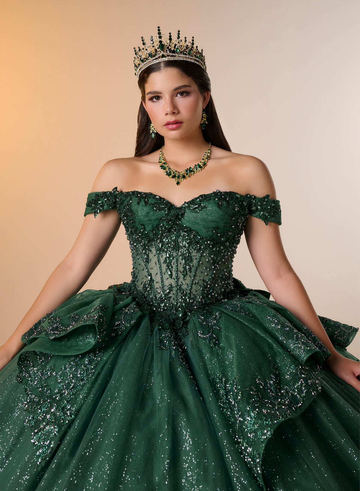 A woman wearing an ornate off-the-shoulder emerald green ball gown with sparkling beadwork and a full layered skirt, styled with a matching green crown, necklace, and earrings against a warm beige studio background. / Luxurious off-shoulder ball gown featuring glittering embellishments, layered draped skirt details, and a dramatic cathedral-length train for a breathtaking princess-inspired look.