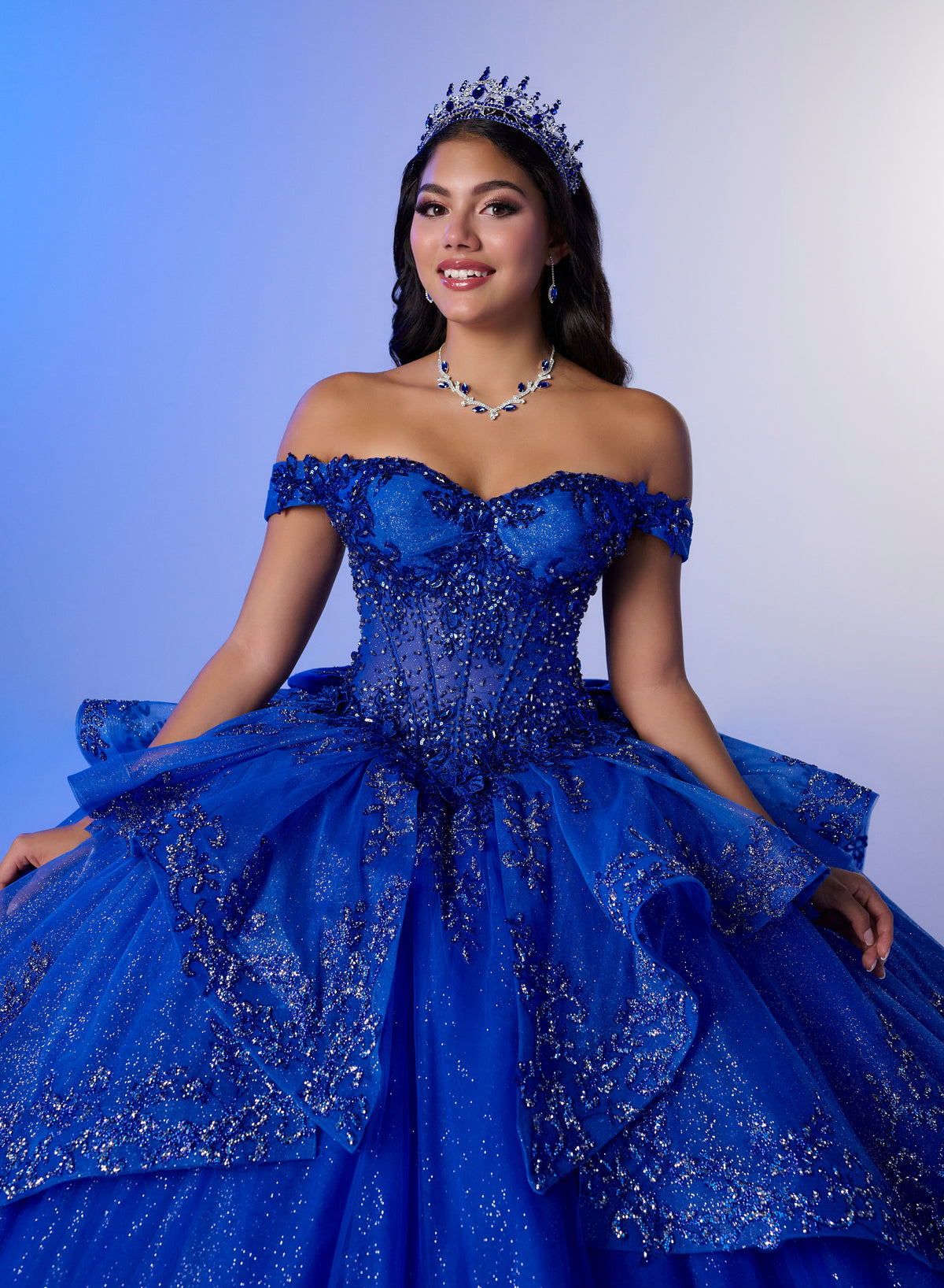 A woman wearing an elaborate royal blue off-the-shoulder ball gown with sparkling embellishments, a matching tiara, and a blue beaded necklace, posed against a blue-to-white gradient studio background. / Luxurious off-shoulder ball gown featuring glittering embellishments, layered draped skirt details, and a dramatic cathedral-length train for a breathtaking princess-inspired look.