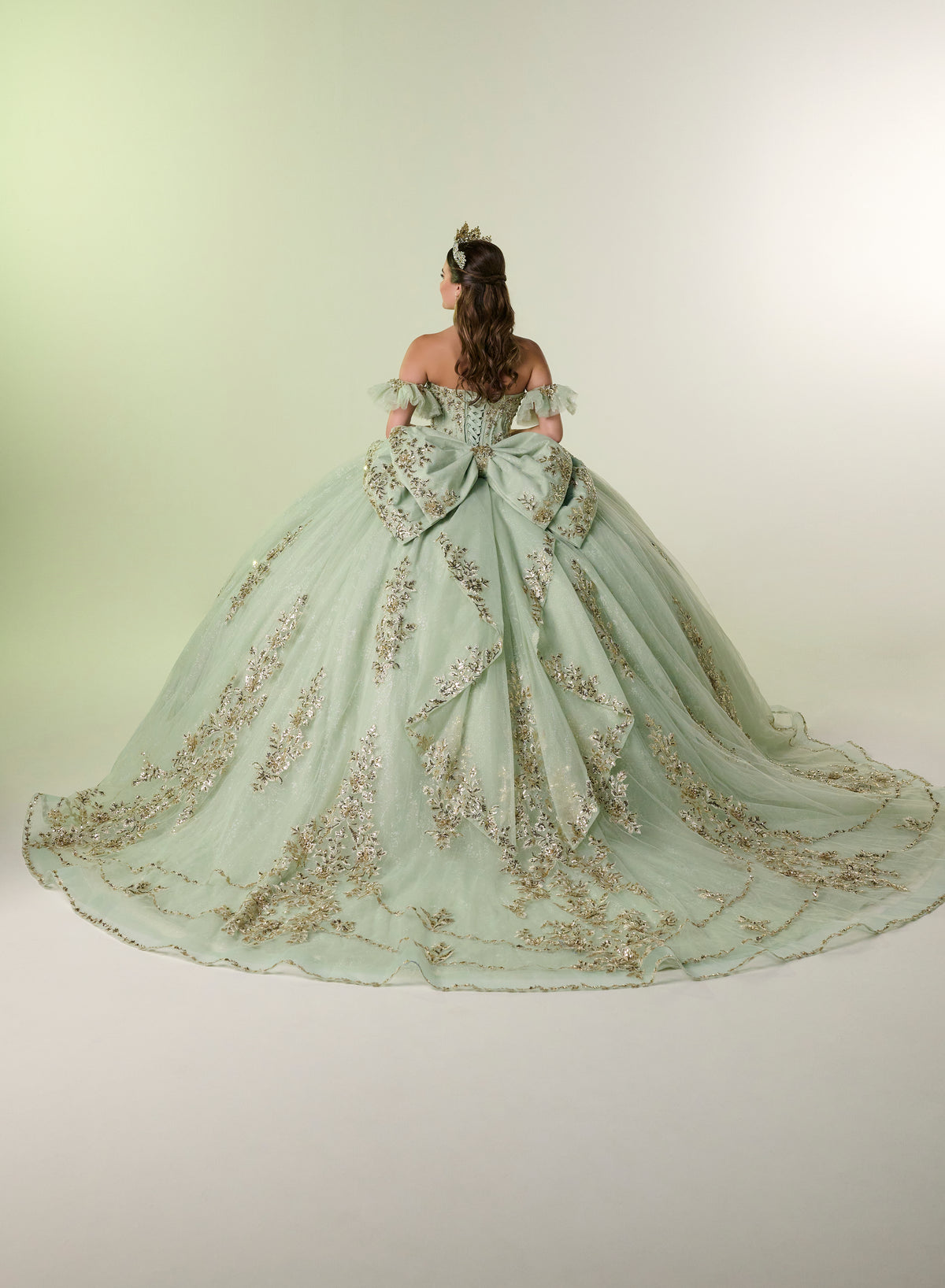 A woman viewed from behind wearing an elaborate off-the-shoulder ball gown in pale mint green with extensive gold floral embroidery and a very large bow at the back, photographed against a plain light studio backdrop. / Elegant off-shoulder ball gown adorned with intricate sparkling appliqués, a structured sweetheart bodice, a voluminous skirt, and a dramatic oversized bow with a sweeping train.