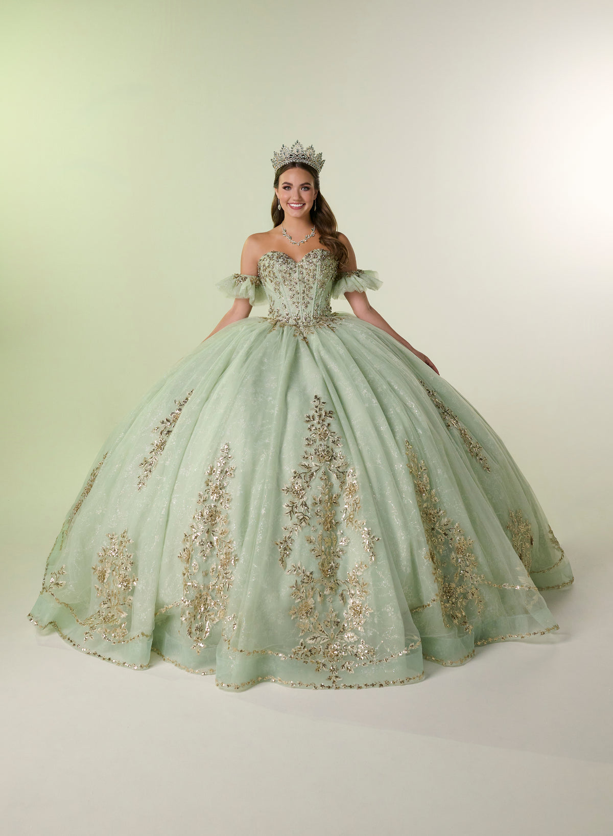 A woman modeling a voluminous mint green ball gown with gold embroidered details, a matching tiara, and a necklace, standing against a light beige studio background. / Elegant off-shoulder ball gown adorned with intricate sparkling appliqués, a structured sweetheart bodice, a voluminous skirt, and a dramatic oversized bow with a sweeping train.