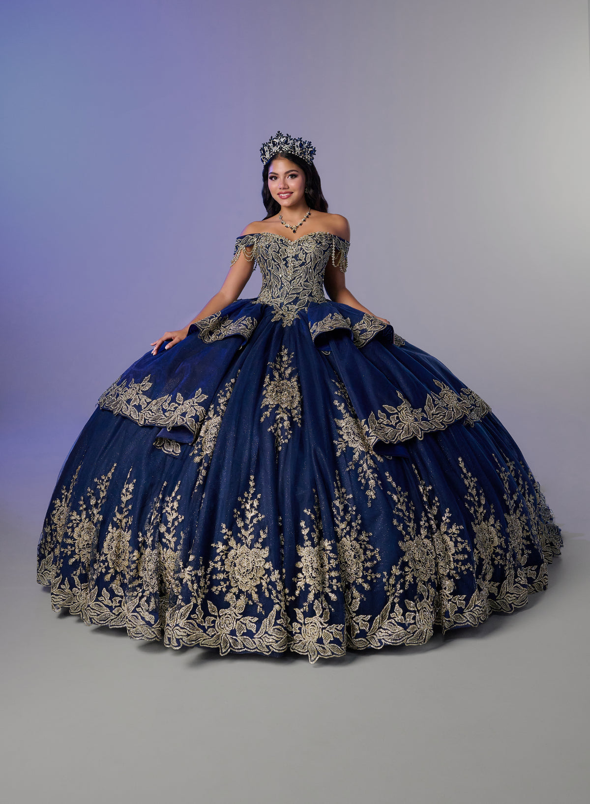 A woman wearing an elaborate off-the-shoulder royal blue quinceañera gown with extensive gold floral embroidery, a matching crown, and a voluminous ball gown skirt, posed against a studio backdrop with a purple-to-gray gradient. / Opulent off-shoulder ball gown featuring intricate embroidered appliqués, dramatic tiered draping, a structured corset bodice, and a sweeping cathedral-length train.