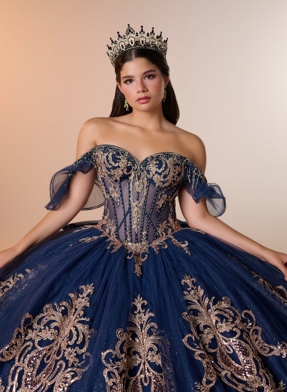 A woman wearing an elaborate off-the-shoulder ball gown in deep navy blue with ornate gold embroidery, a matching crown, and dangling earrings, posed against a warm beige studio background. / Regal off-shoulder ball gown adorned with intricate embroidered appliqués, a structured corset bodice, and a voluminous skirt with an elegant sweeping train.