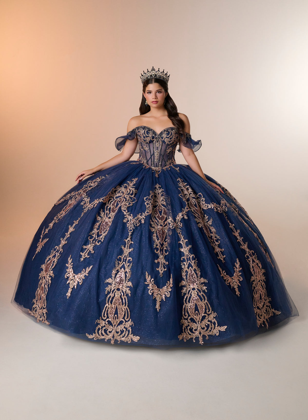 A woman wearing a voluminous royal blue ball gown with ornate gold embroidery, a corset-style bodice, and sheer off-the-shoulder sleeves, posed against a warm beige studio background. She is also wearing a small tiara. / Regal off-shoulder ball gown adorned with intricate embroidered appliqués, a structured corset bodice, and a voluminous skirt with an elegant sweeping train.