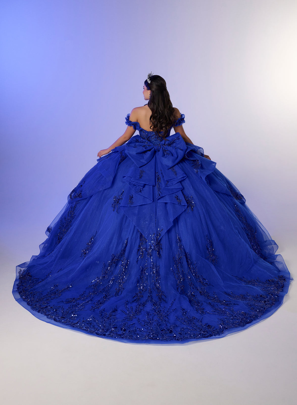 A woman wearing a voluminous royal blue ball gown with intricate beadwork and a large bow at the back, photographed against a soft blue-to-white studio backdrop. / Elegant off-shoulder ball gown featuring sparkling embellished appliqués, a structured corset bodice, dramatic layered skirt details, and a voluminous princess silhouette.