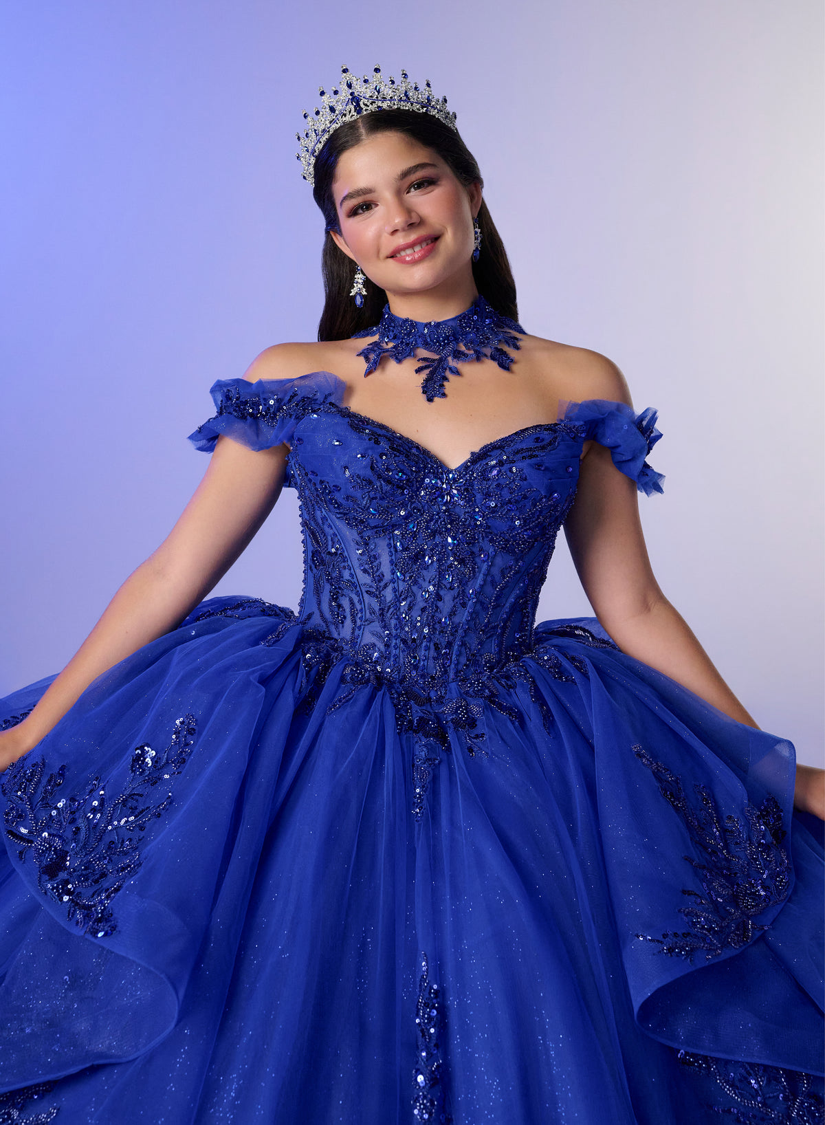 A woman wearing a royal blue off-the-shoulder ball gown with intricate beadwork, a matching blue choker, and a silver tiara, posing against a light blue studio background. / Elegant off-shoulder ball gown featuring sparkling embellished appliqués, a structured corset bodice, dramatic layered skirt details, and a voluminous princess silhouette.