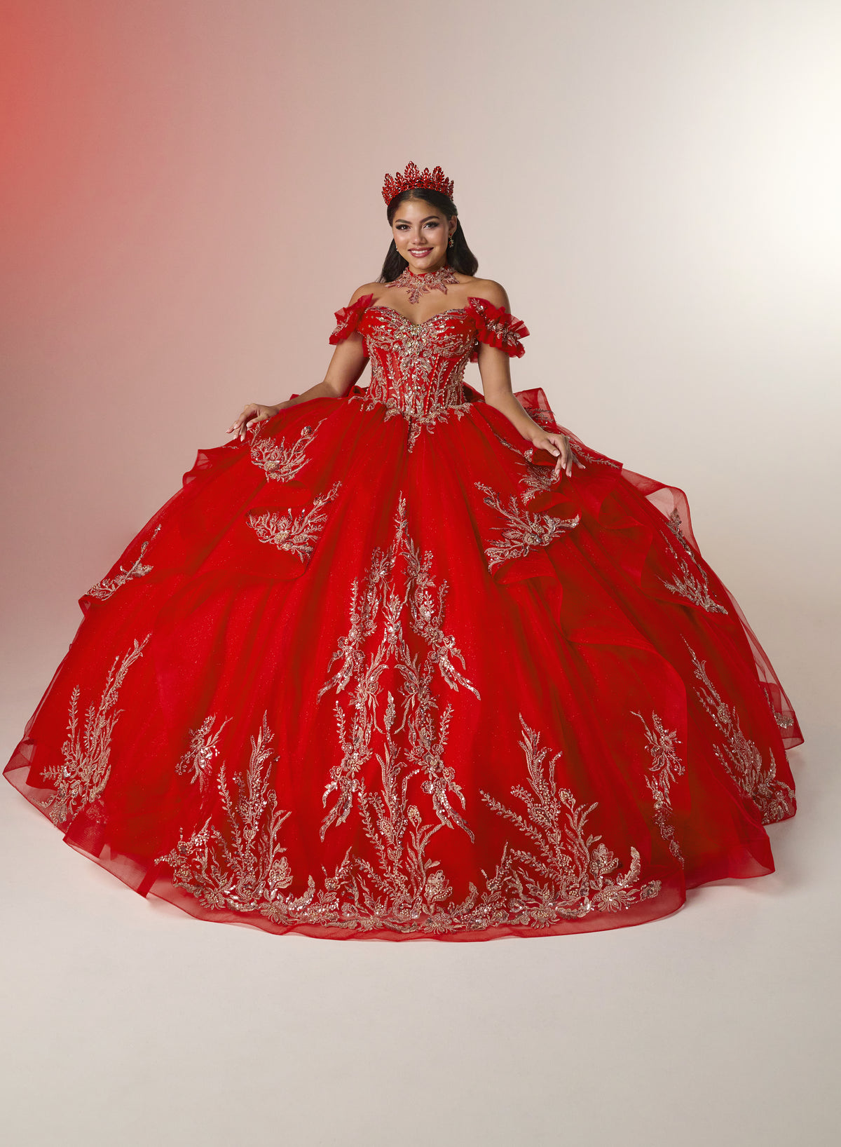 A woman wearing a voluminous red ball gown with silver floral embroidery, off-the-shoulder ruffle sleeves, a jeweled red crown, and a matching red necklace, posed against a warm off-white studio background. / Elegant off-shoulder ball gown featuring sparkling embellished appliqués, a structured corset bodice, dramatic layered skirt details, and a voluminous princess silhouette.