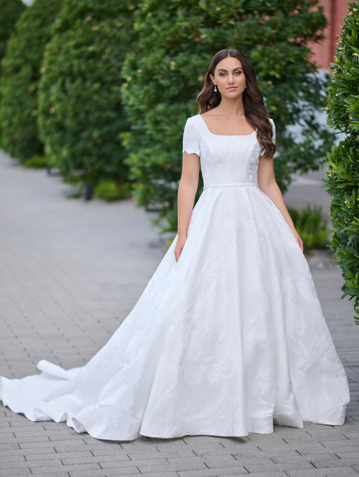 Woman in a white wedding dress standing outdoors with greenery in the background / A-line style in jacquard with soft square neckline and short sleeves finished with scallop shape. Covered buttons at the back closure, buttons continuing all the way down to hemline along the train.