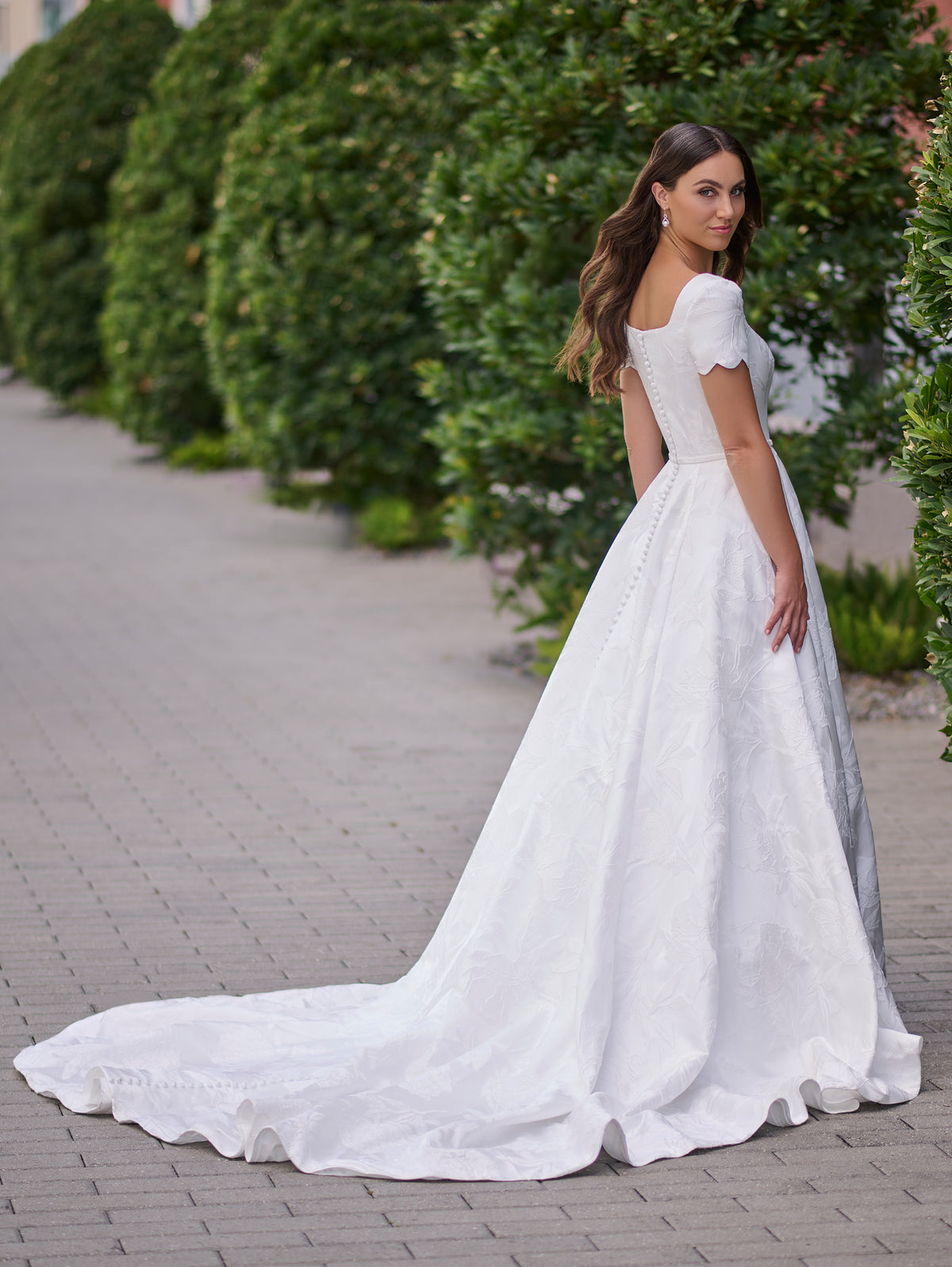 A woman wearing a white off-the-shoulder A-line wedding gown with a long train, standing on a paved walkway with green hedges in the background. / A-line style in jacquard with soft square neckline and short sleeves finished with scallop shape. Covered buttons at the back closure, buttons continuing all the way down to hemline along the train.