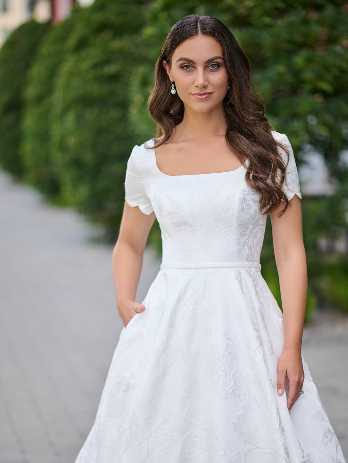 A woman wearing a white A-line wedding dress with short puff sleeves, a square neckline, and a textured lace overlay, standing outdoors on a paved path with a green hedge in the background. / A-line style in jacquard with soft square neckline and short sleeves finished with scallop shape. Covered buttons at the back closure, buttons continuing all the way down to hemline along the train.