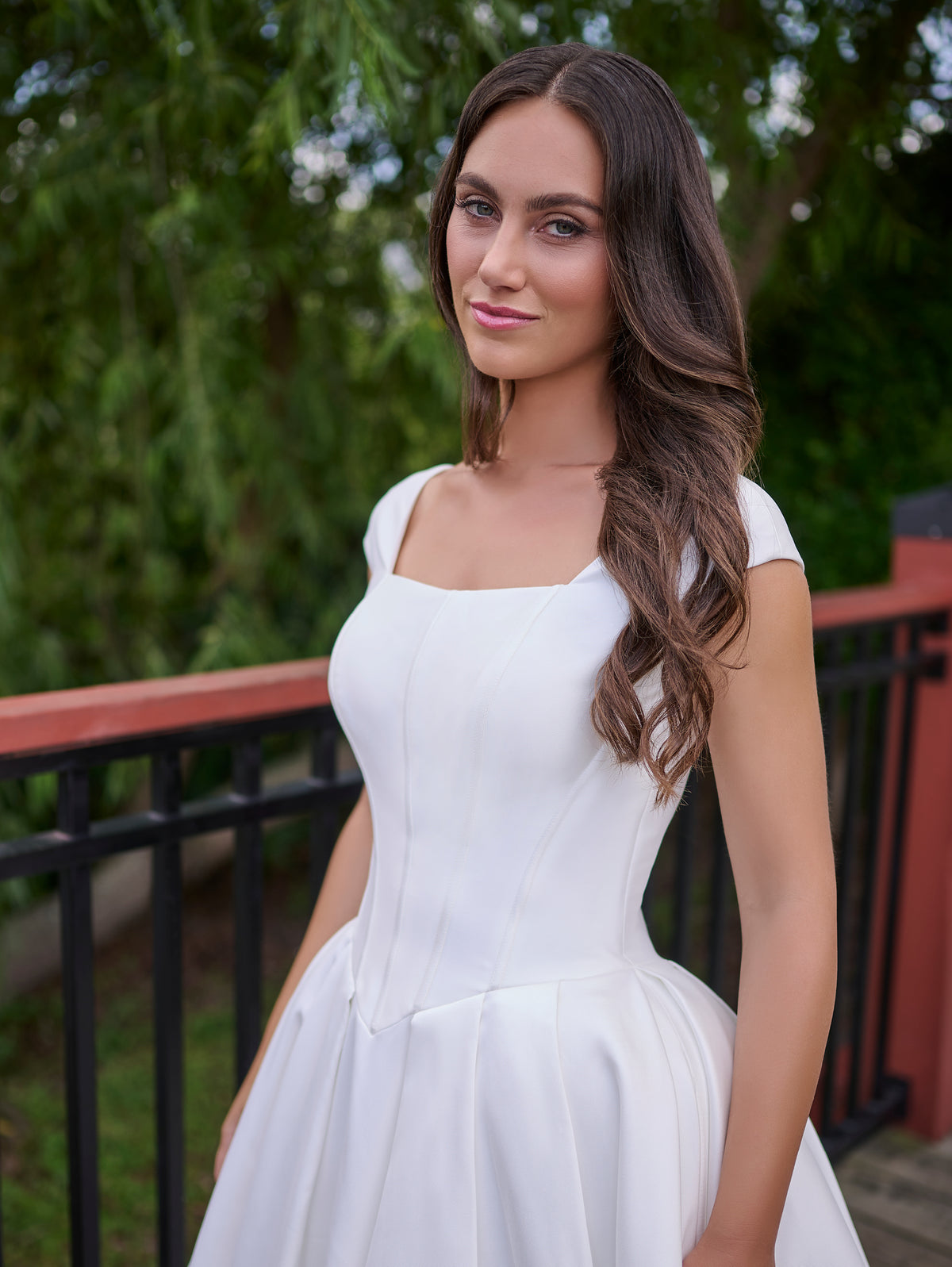 A woman wearing a white sleeveless fit-and-flare dress with a square neckline and full skirt, standing outdoors in front of a black railing and green foliage. / Clean mikado ball gown with structured bodice and princess skirt with pleats. The drop waist bodice with exposed boning has square neckline and cup sleeves. Covered buttons all the way to hemline on CB closure finishes the look.