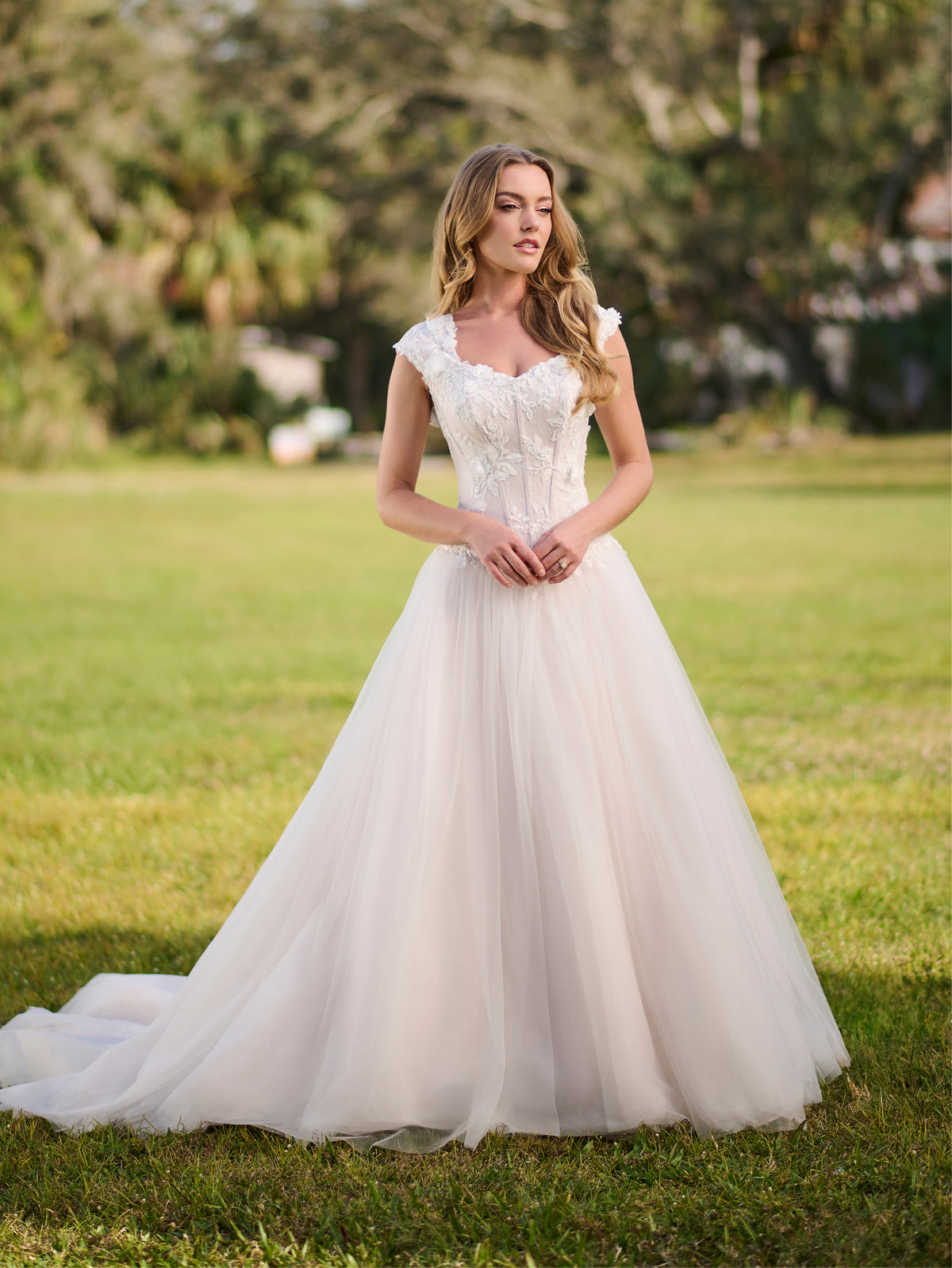 Woman in a white wedding dress standing on grass with trees in the background / Cap sleeve Basque waist tulle ballgown with corsetry lines and 3D flower lace appliques.