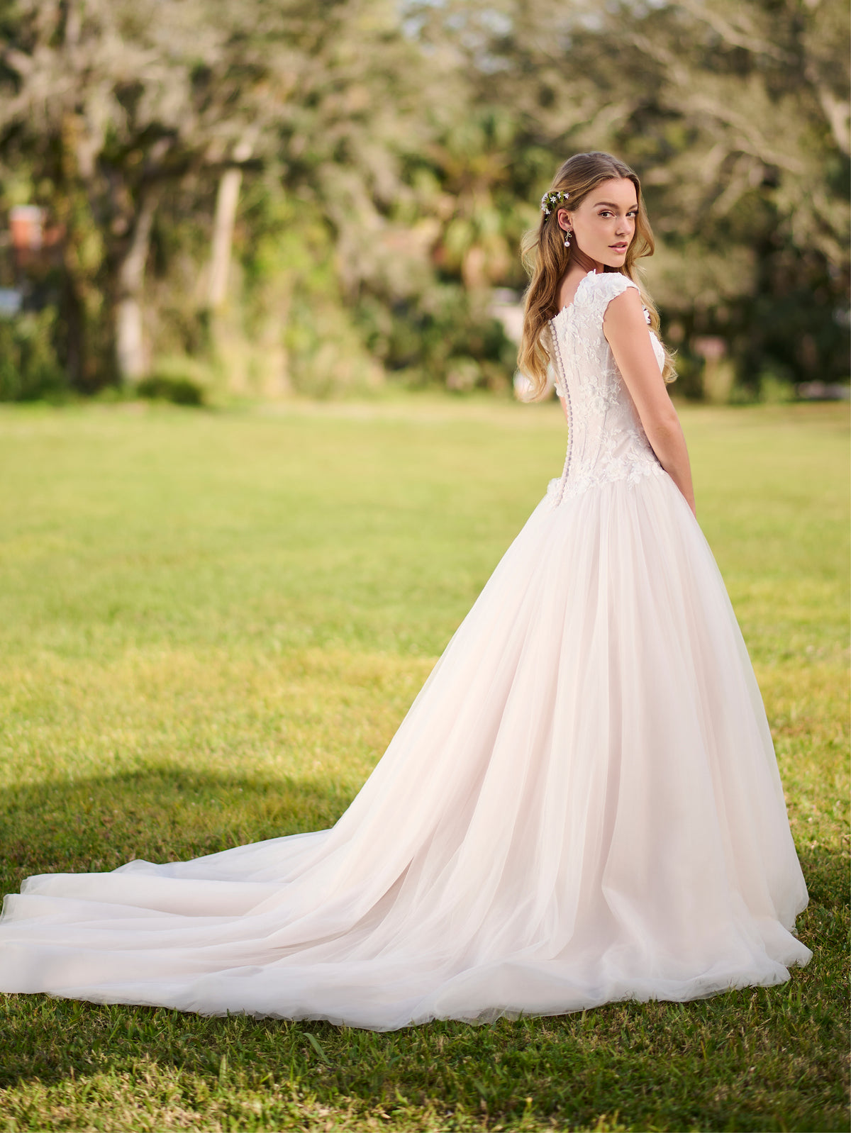 Woman in a white wedding dress standing on grass with trees in the background / Cap sleeve Basque waist tulle ballgown with corsetry lines and 3D flower lace appliques.
