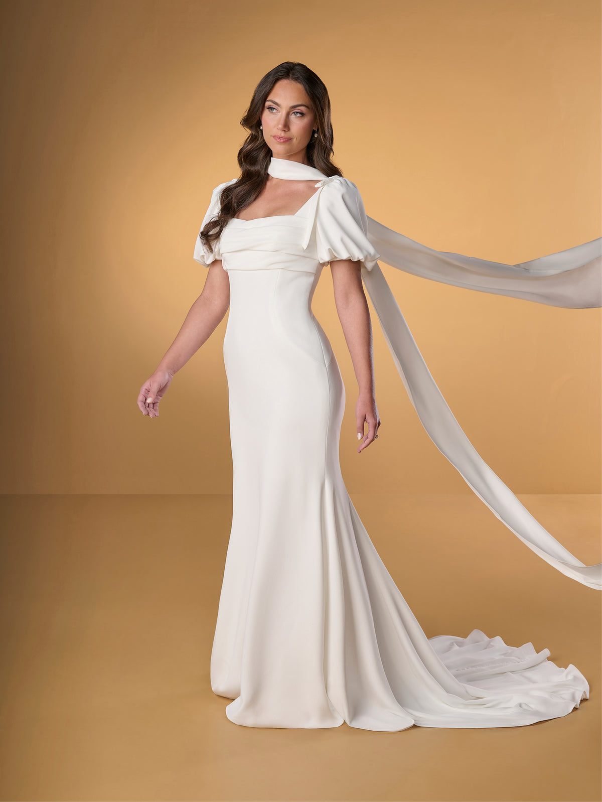 A woman modeling a floor-length ivory wedding gown with a high draped neckline, short flutter sleeves, and a long flowing train against a warm tan studio backdrop. / Slim crepe dress with square neckline and puffy short sleeves. Buttons at the back along zipper closure and all the way down on train.