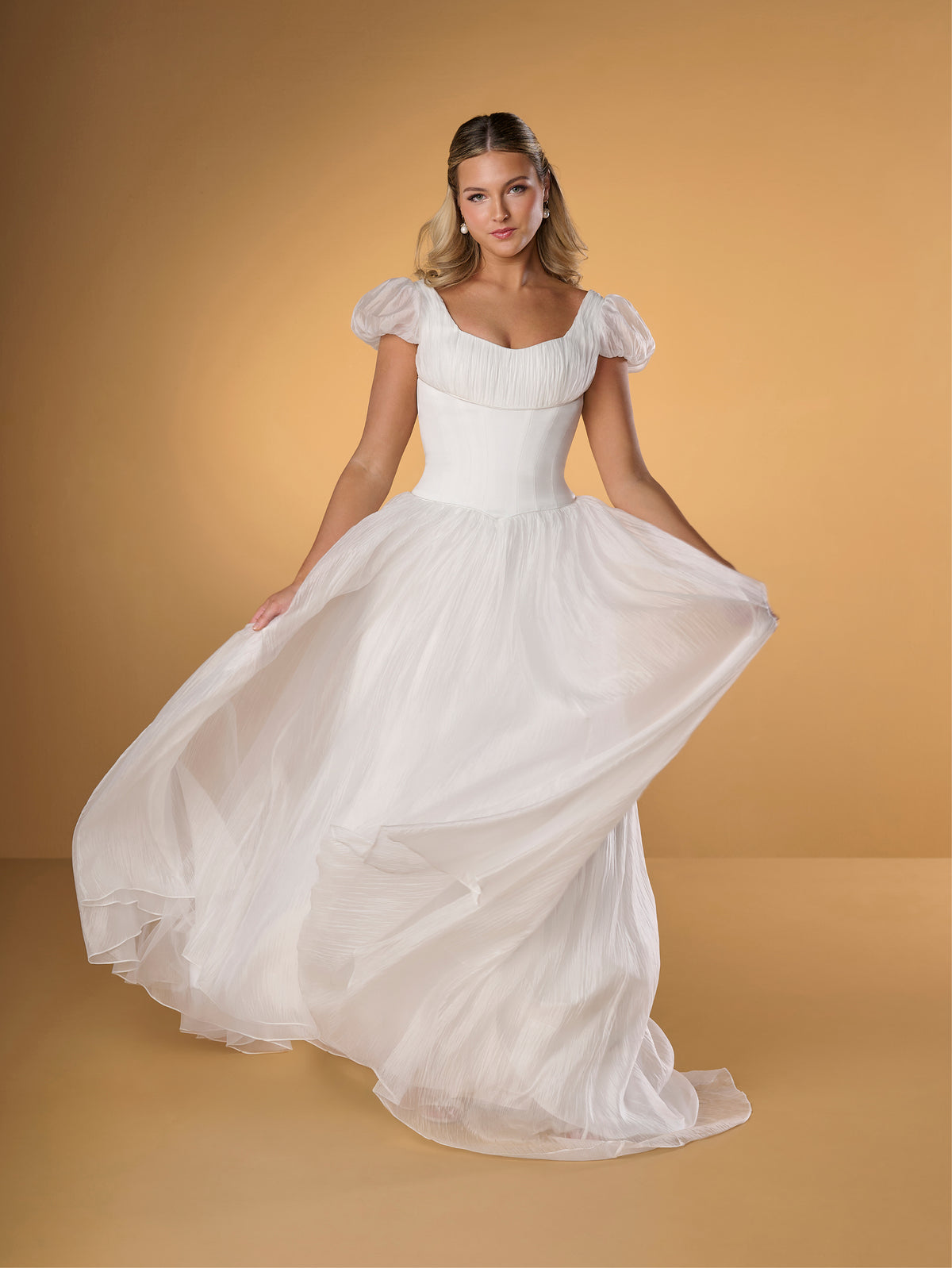 A woman modeling a white off-the-shoulder ballgown with a fitted bodice and a voluminous flowing skirt, posed against a warm tan studio backdrop. / A-line gown in crinkled organza and satin midriff area on bodice only. Short puffy sleeves. Covered buttons along zipper closure at the back dress.