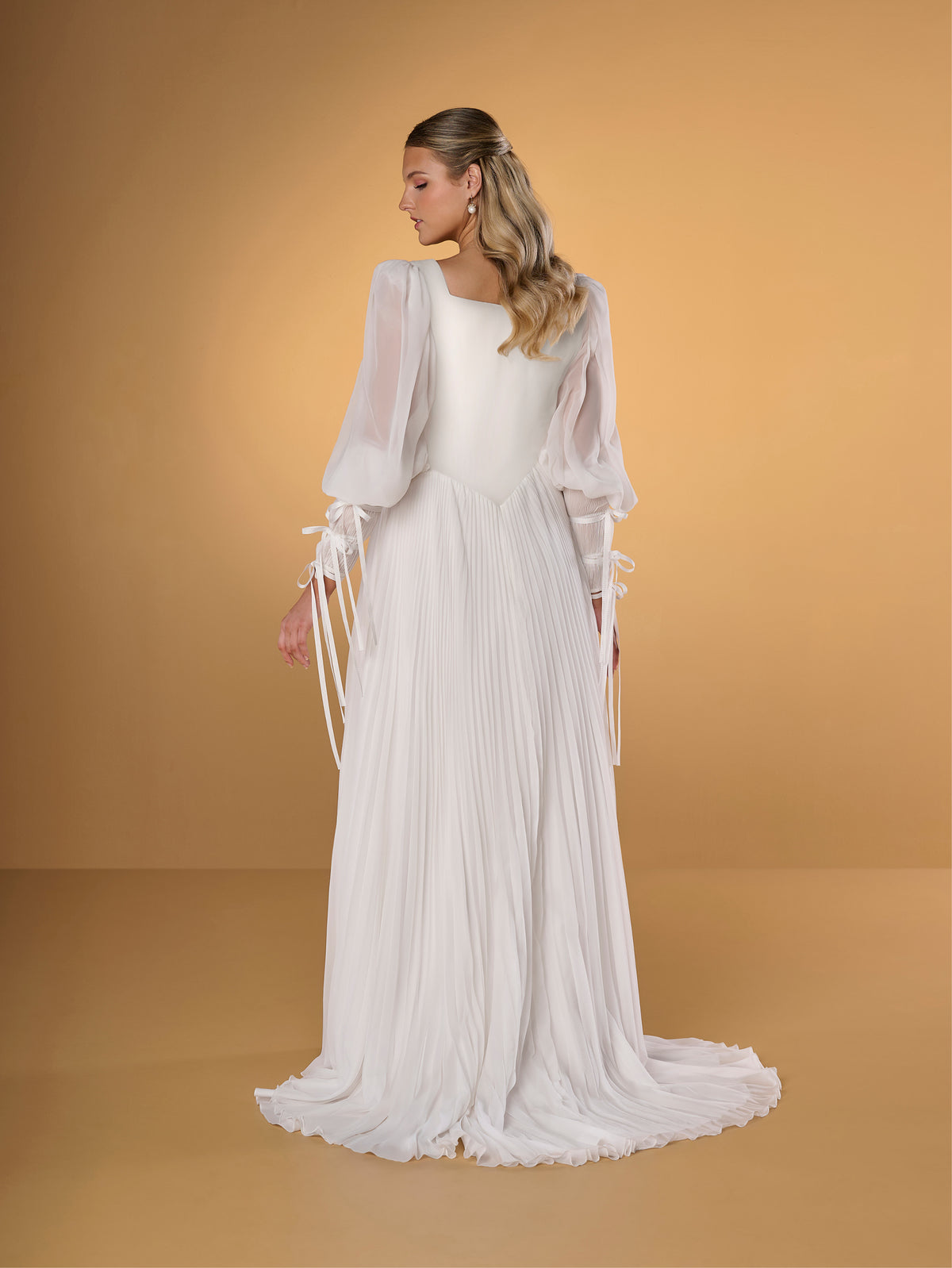 A woman modeling a long white dress with sheer balloon sleeves, tie details at the cuffs, and a pleated skirt, posed against a warm tan studio backdrop. / A-line style in chiffon with square neckline shape, permanently pleated chiffon on skirt area only. Puffy sleeves fitted on long cuffs, decorated with narrow ribbons.