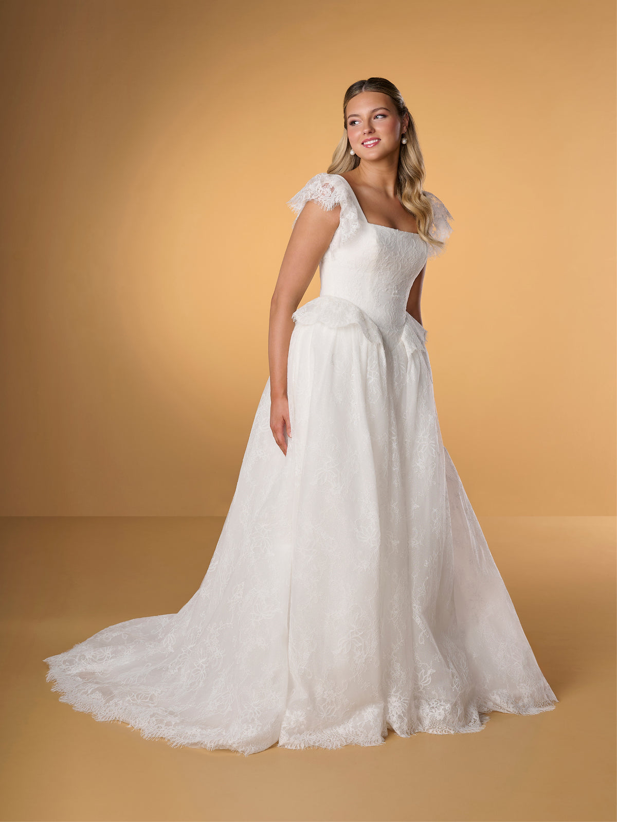 Woman wearing a white lace wedding dress against a yellow background / Lace ball gown with square neckline, lace peplums and flatter sleeves. Covered buttons along zipper back closure.