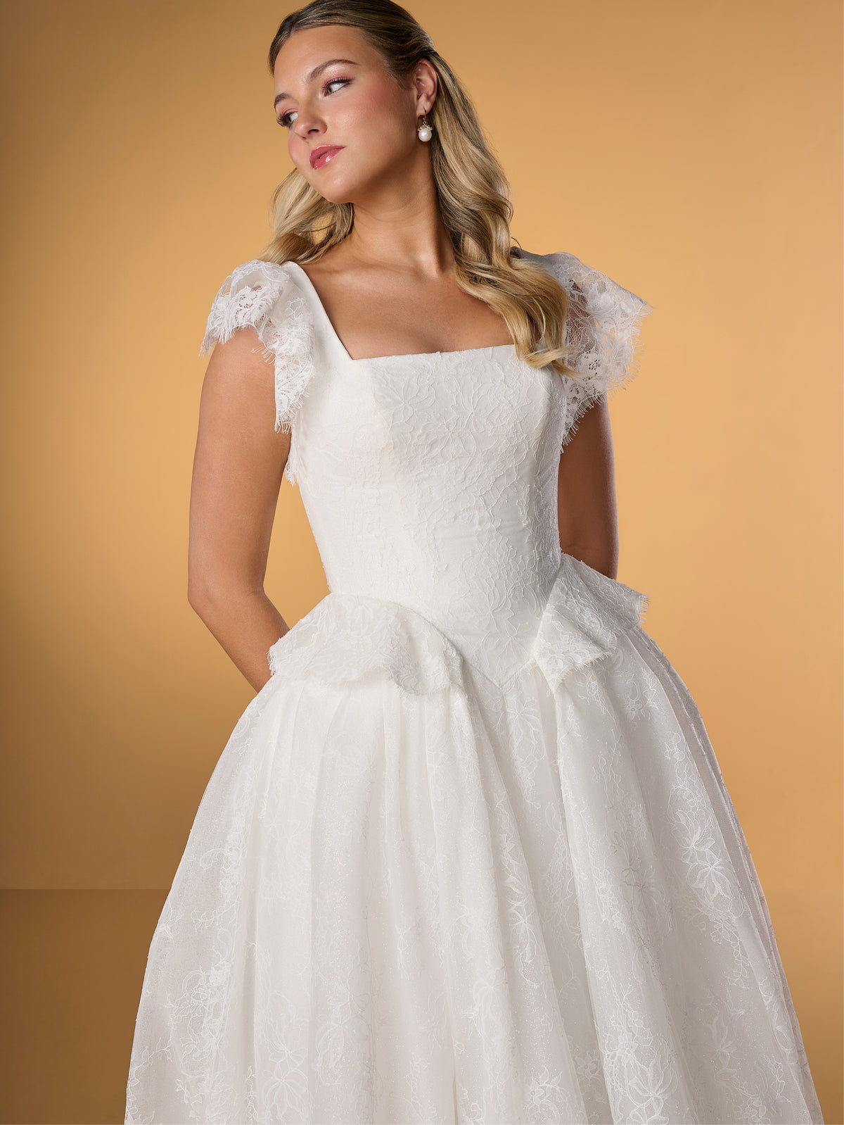 Woman wearing a white wedding dress with lace cap sleeves against a yellow background / Lace ball gown with square neckline, lace peplums and flatter sleeves. Covered buttons along zipper back closure.