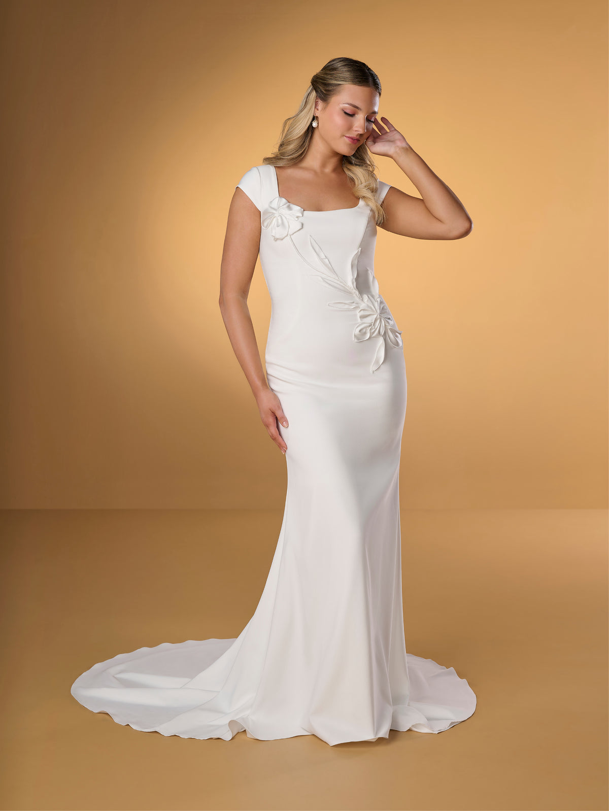 Woman wearing a white wedding dress with floral details on a beige background / Slim crepe style with square neckline and cup sleeves, decorated with 3D motifs in soft crinkle organza fabric. Covered buttons along zipper closure at the back.