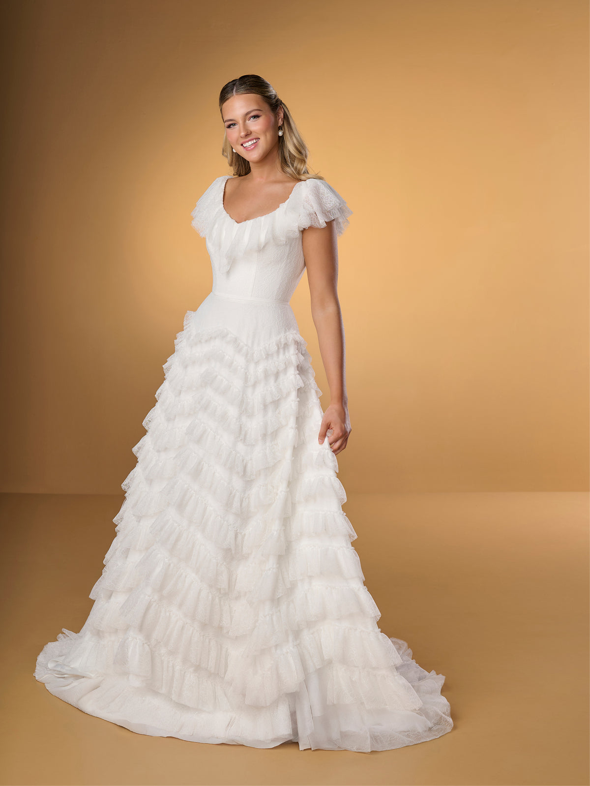 Woman wearing a white wedding dress with ruffled details against a gold background / A-line style in delicate lace with ruffles around scooped neckline and all over the skirt. Invisible zipper closure at the back.