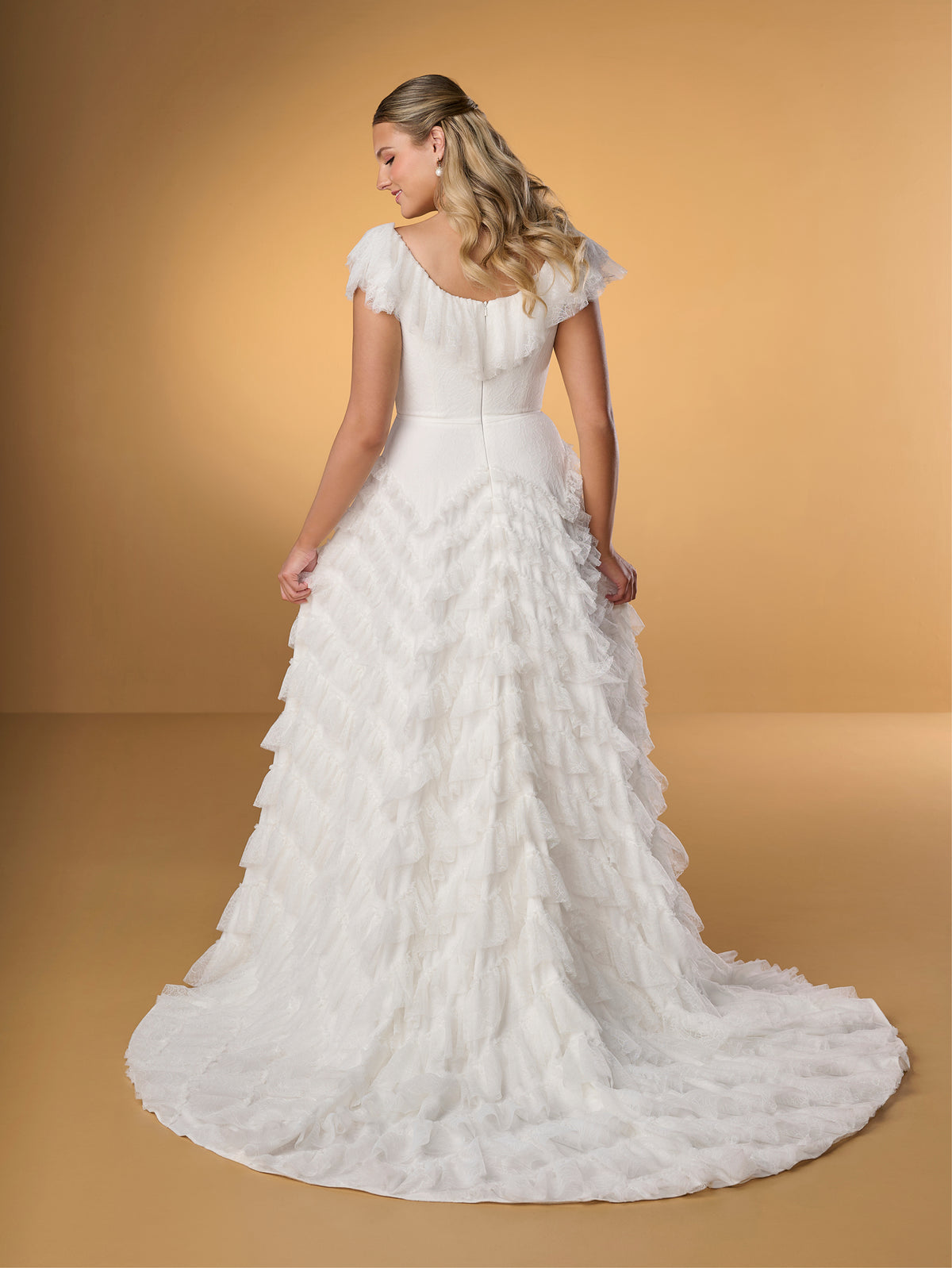 Woman wearing a white wedding dress with ruffled details on a beige background / A-line style in delicate lace with ruffles around scooped neckline and all over the skirt. Invisible zipper closure at the back.