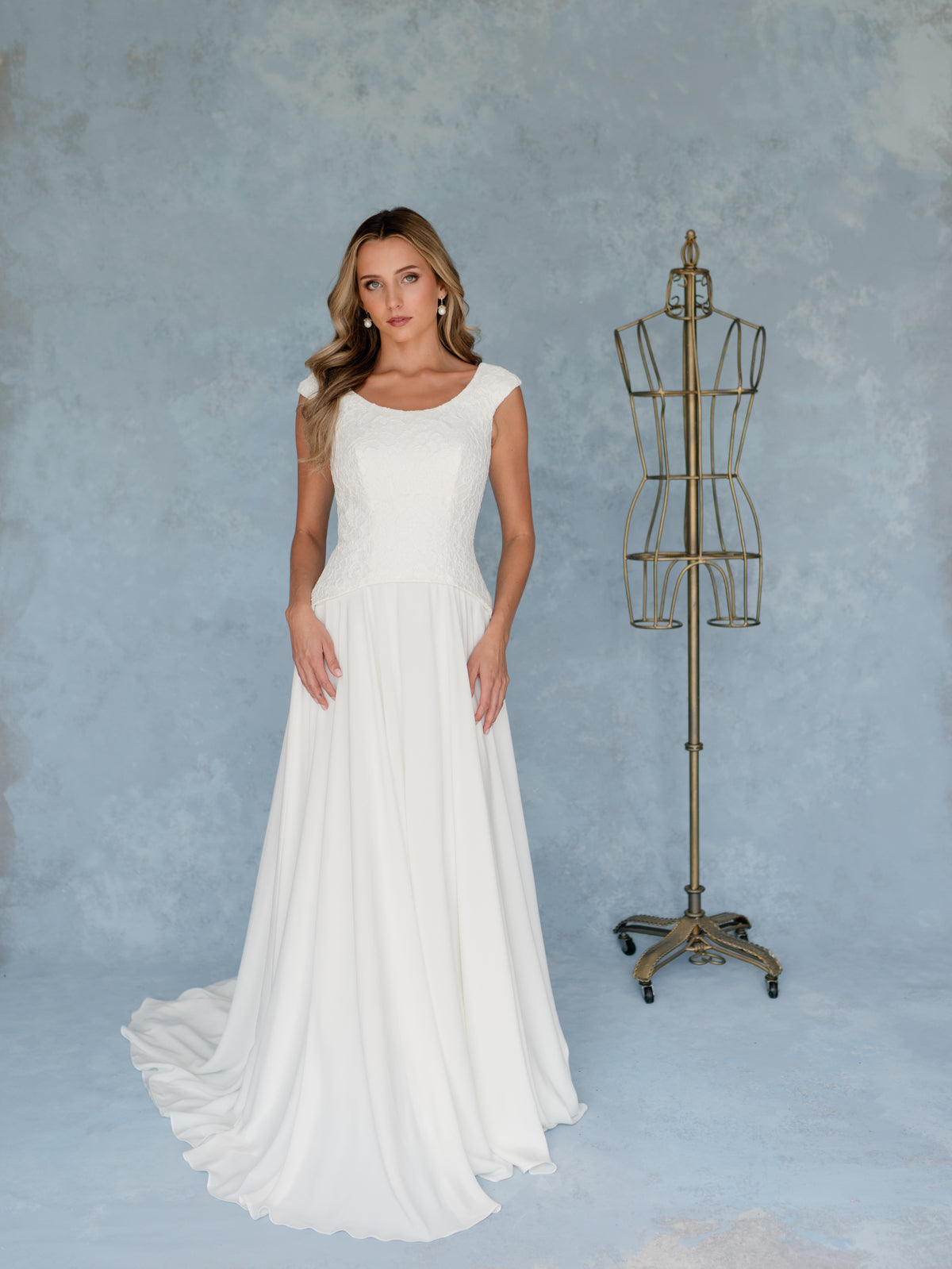 A woman modeling a sleeveless white wedding gown with a fitted bodice and flowing skirt, standing beside a vintage-style metal dress form against a textured light blue studio backdrop. / A-line style - mock 2 piece- with lace bodice and chiffon skirt. Scooped neckline, zipper/buttons back closure.