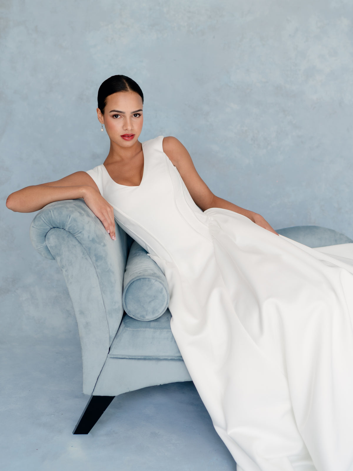 A woman reclining on a light blue velvet chaise lounge against a textured light blue studio backdrop, wearing a sleeveless white wedding dress with a deep V-neck and flowing skirt. / Sleeveless A-line style in lux satin with V-neck and zipper/buttons back closure - button all the way to hemline.