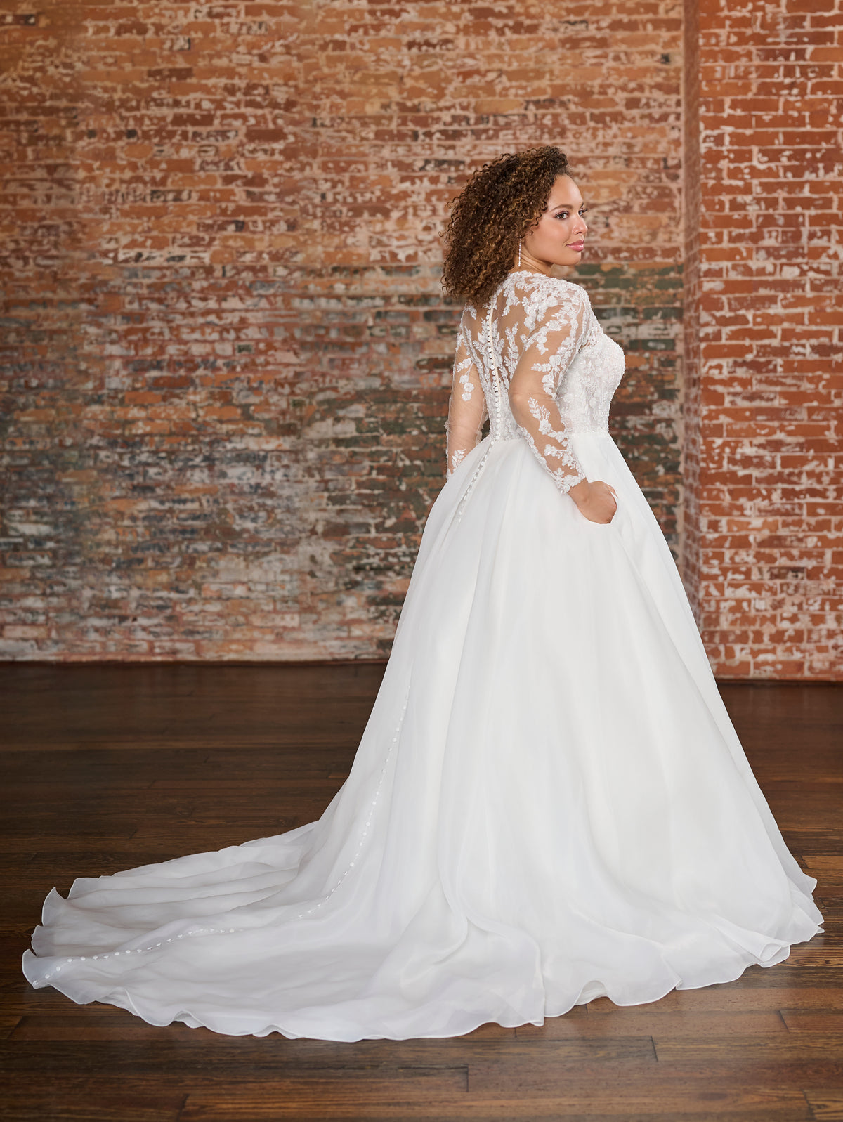 A woman wearing a white wedding dress with lace long sleeves and a full tulle skirt, standing in a room with a brick wall and wooden floor. / This beautiful, A-line ballgown has a semi sheer bodice that is made of layers of beaded tulle, lace and plain tulle to create the perfect amount of coverage. Boning has been placed to perfectly support the body through the bodice. The flattering square neckline is complimented by long, sheer sleeves and an illusion back bodice. The skirt begins at the natural