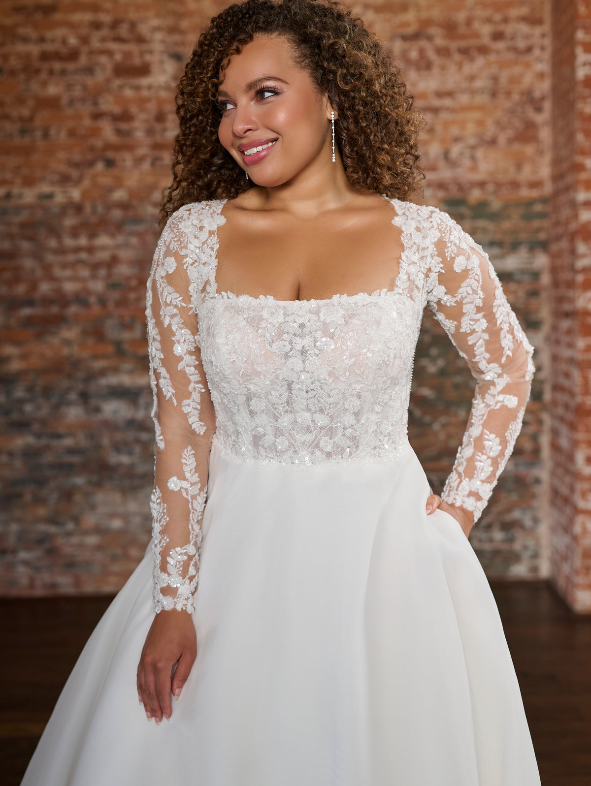 A woman wearing a white wedding dress with a lace bodice, sheer long sleeves, and a full skirt, standing in front of a rustic brick wall. / This beautiful, A-line ballgown has a semi sheer bodice that is made of layers of beaded tulle, lace and plain tulle to create the perfect amount of coverage. Boning has been placed to perfectly support the body through the bodice. The flattering square neckline is complimented by long, sheer sleeves and an illusion back bodice. The skirt begins at the natural