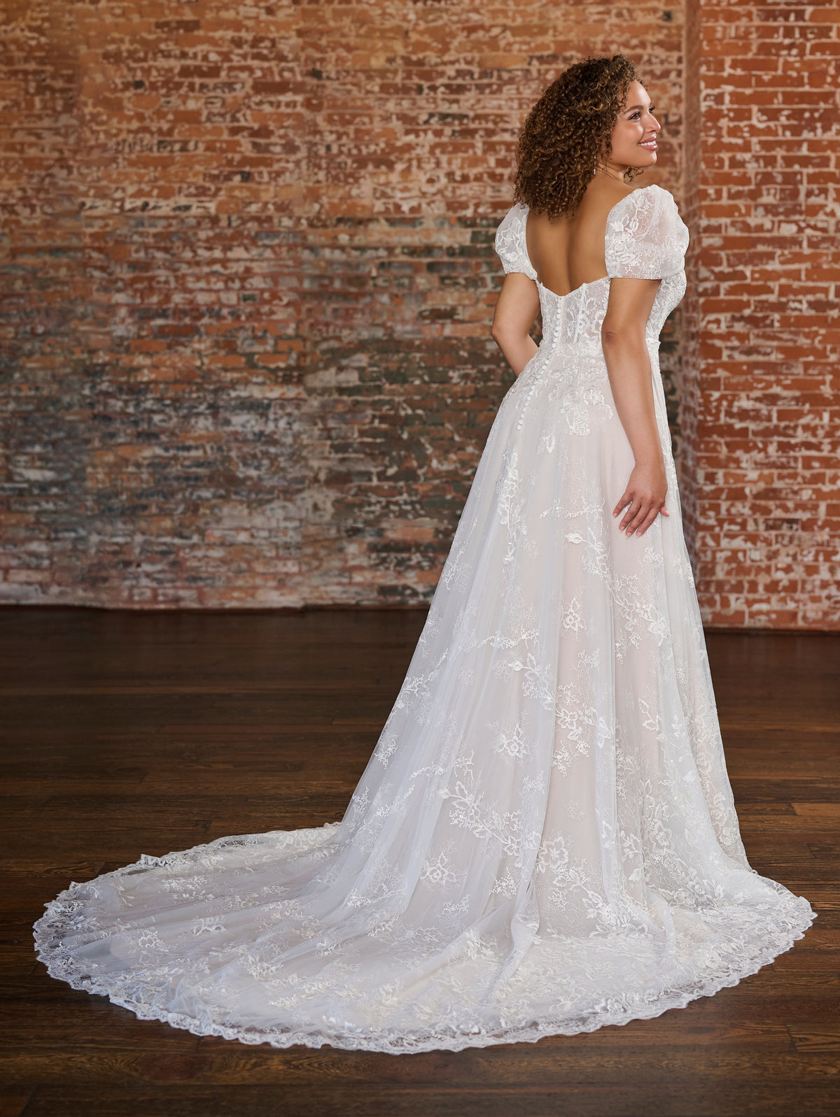 A woman wearing a white lace wedding gown with short puff sleeves and a long train, photographed from the back in front of a rustic brick wall and dark wood floor. / Soft romantic A-line dress in airy all over lace with sweetheart neckline and short balloon sleeves. Semi sheer bodice decorated with beautiful floral lace. Covered buttons at the back closure, long train.