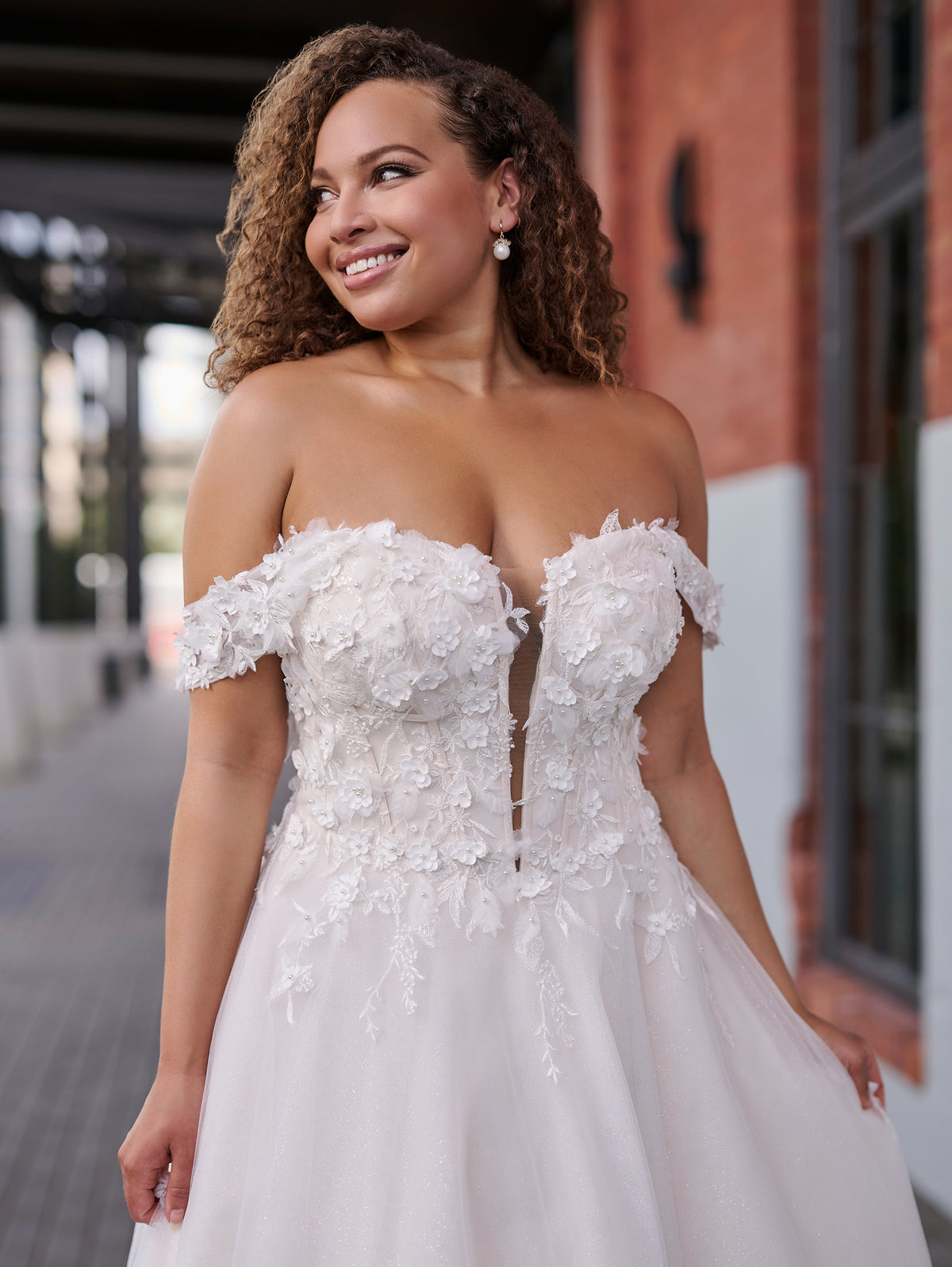 A woman wearing a strapless white wedding gown with floral lace applique and a tulle skirt, photographed outdoors in front of a brick building. / Soft A-line gown in tulle with 3D lace motifs decorating bodice with sweetheart neckline and deep illusion v on center front. Off-shoulder straps. Long train.