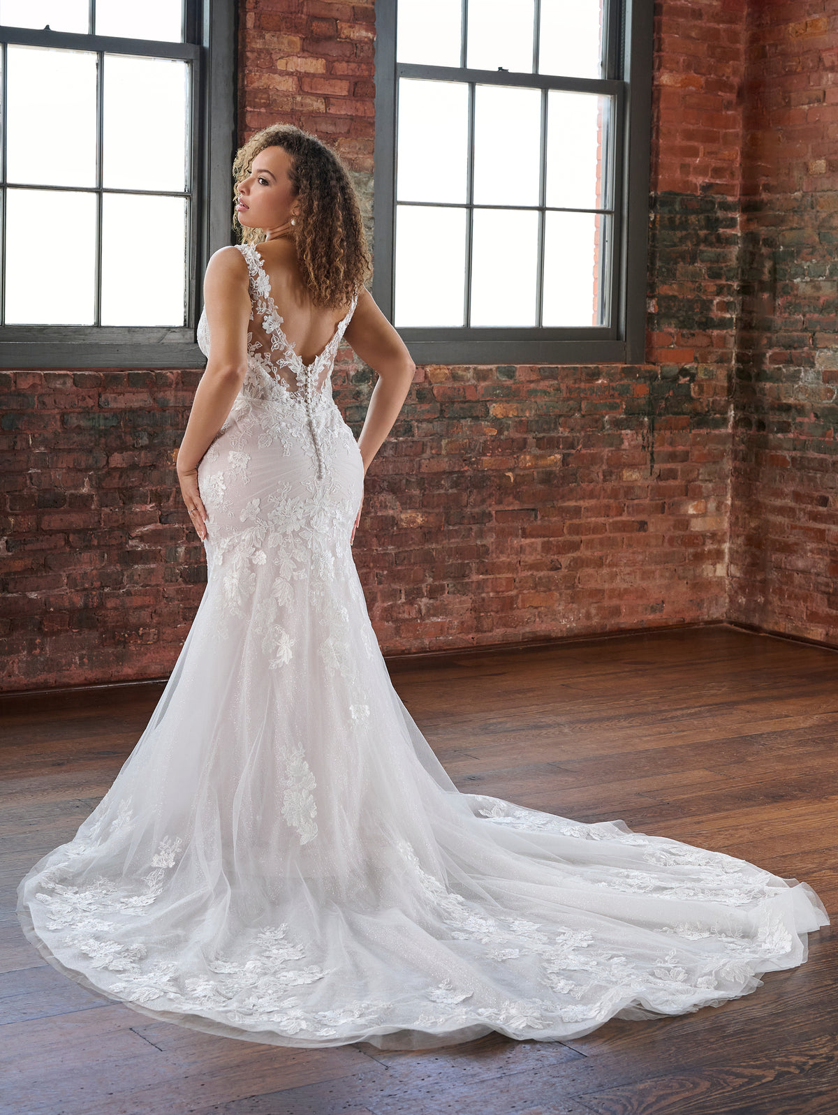 A woman wearing a white lace mermaid-style wedding gown with a long train, photographed indoors in a brick loft with large windows. / This slim and sexy gown has placed lace appliques that sit on a layer of tulle that is placed over sparkle tulle. The semi sheer bodice leads to sheer straps and a sheer back bodice. There are flattering pleats at the back and a long sheer train.