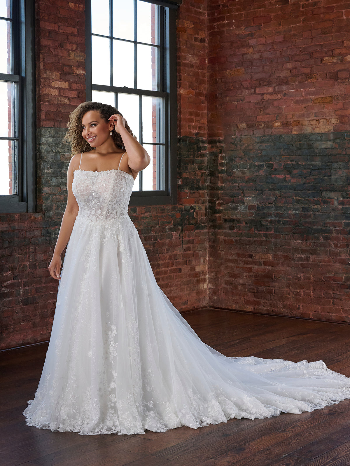 A woman modeling a strapless white wedding gown with a fitted lace bodice and a long sheer tulle train, standing in a brick warehouse room with large windows. / Soft A-line style with square neckline, this style features floral lace motifs with 3D petals.