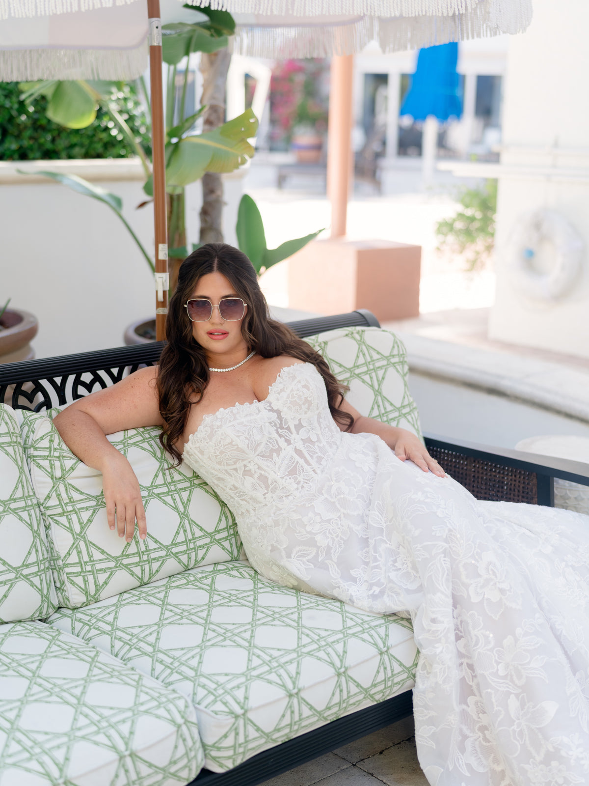 A woman reclining on a patterned outdoor sofa wearing a strapless white lace wedding gown and sunglasses in a bright patio setting. / Lace A-line style with sweetheart neckline and lace up back closure.