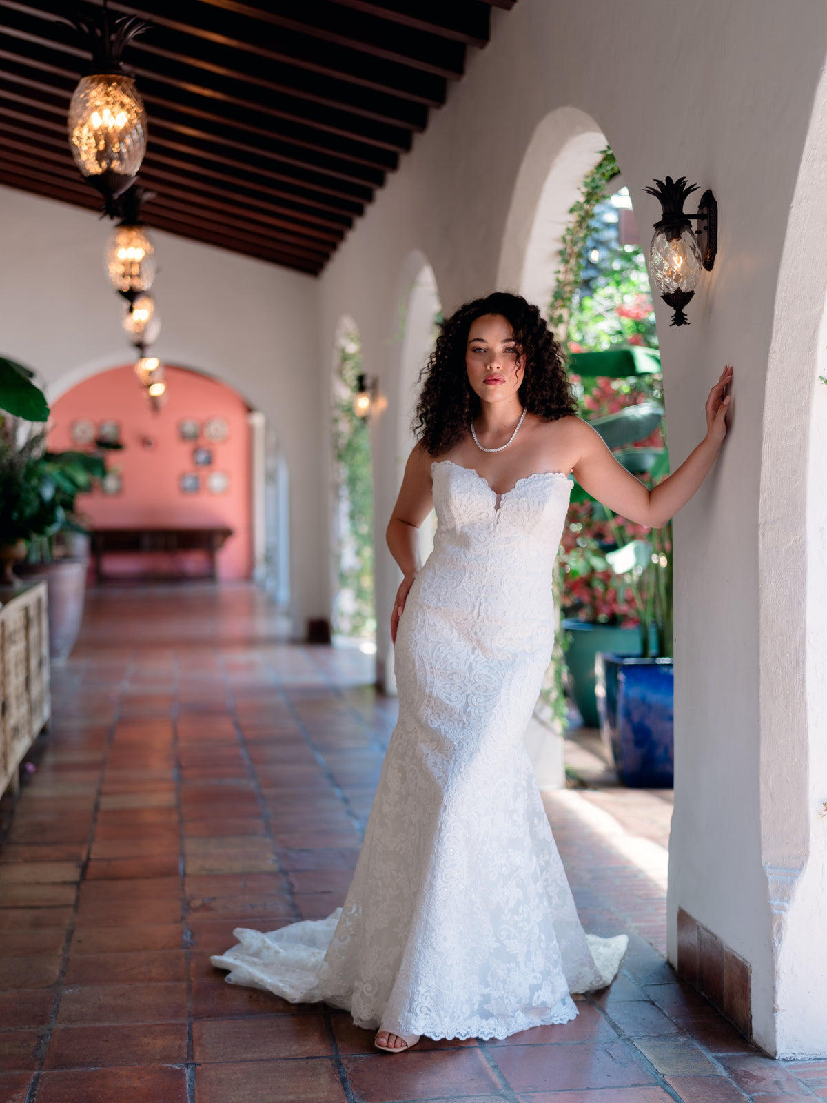 A woman in a strapless white lace wedding gown with a sheer veil and a long flowing train stands in a dimly lit interior. She holds a small tan clutch, and the background includes dark furniture and warm-toned wall panels. / Placed lace slim gown with scallop detail.