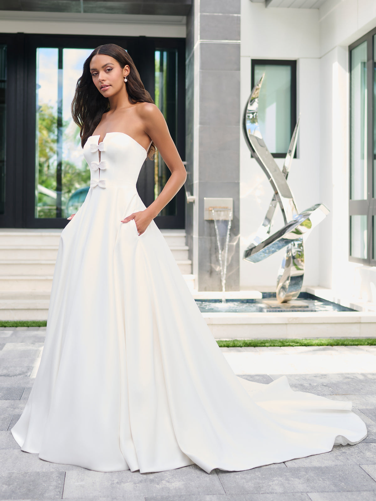 A woman modeling a strapless white wedding gown with a fitted bodice, lace-up front, and full flowing skirt with a long train, standing outdoors in front of a modern building facade. / Luxe matte satin has been used to create this beautiful A-line ballgown. The bodice features a plunging center front neckline that has been adorned with pearl encrusted bows. The full skirt is semi-circle cut and has pockets at the side seam.