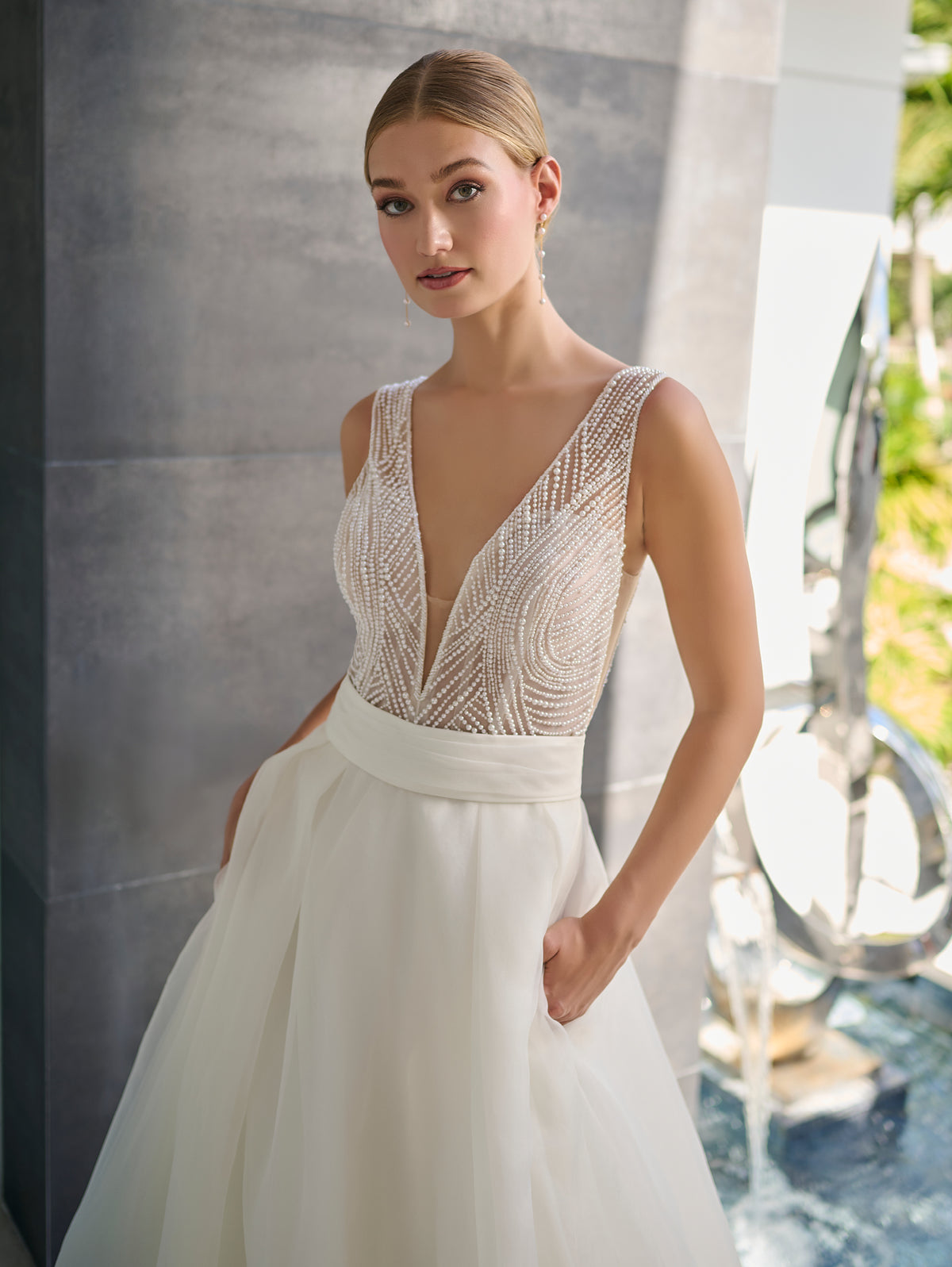 A woman modeling a sleeveless white wedding gown with a deep V-neckline, heavily embellished beaded bodice, and a full tulle skirt, posed against a gray stone wall. / Hand beaded bodice made of strings of various sized pearls with beaded edge finishing. There are sheer inserts at the side and a full illusion back bodice. The skirt is made a matte organza with flat pleats to create soft volume. A soft, pleated organza belt wraps around the waist