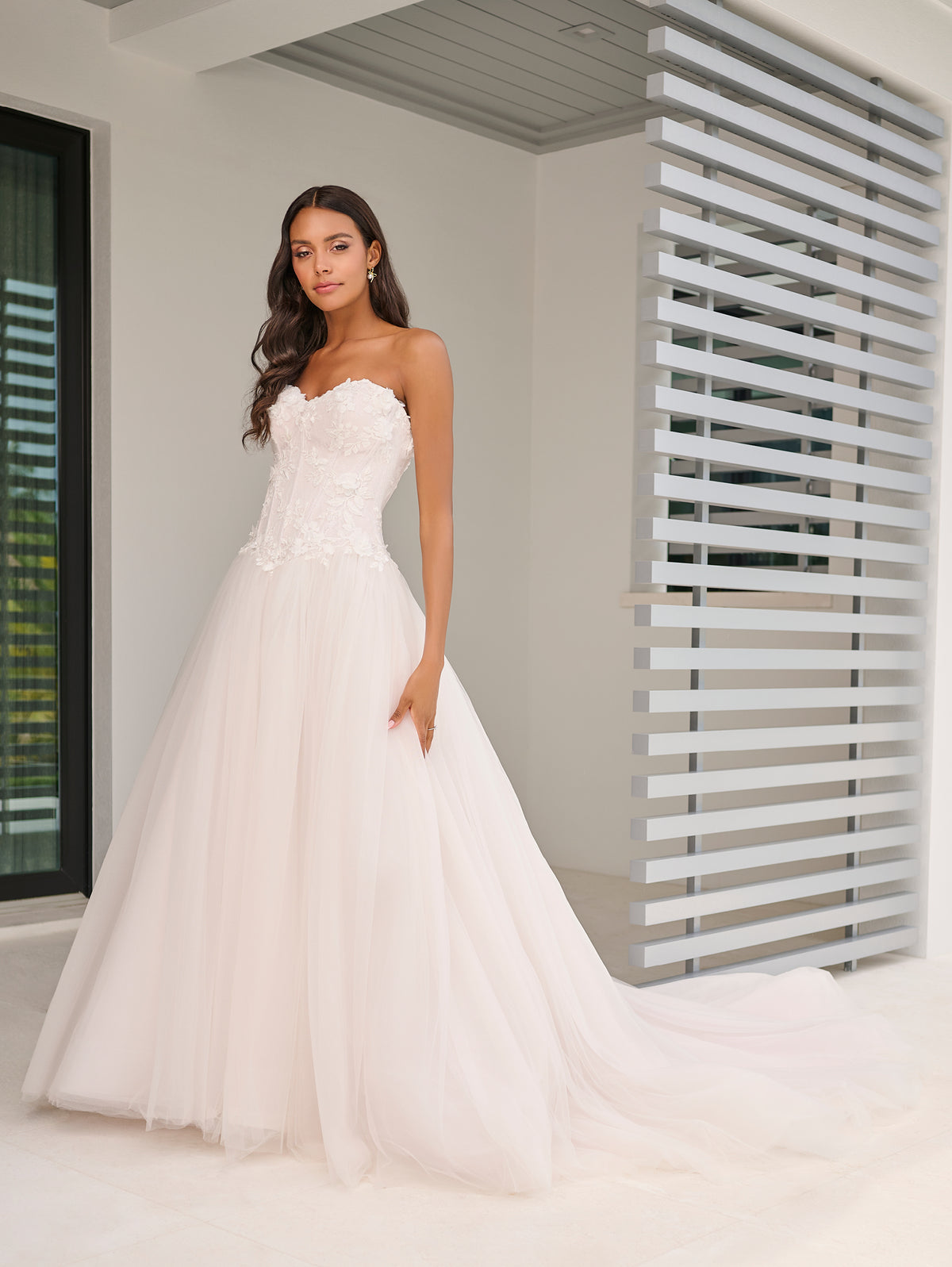 A woman modeling a strapless white wedding gown with a fitted lace bodice and a full tulle skirt with a long train, standing in a bright modern interior with white walls and horizontal slat details. / This dropped waist bodice is made of a stunning 3D floral lace that has been placed over a layer of chantilly lace. The encased boning adds beautiful structure and secure fit. The dramatic skirt is made of several layers of tulles that all work effortlessly together.