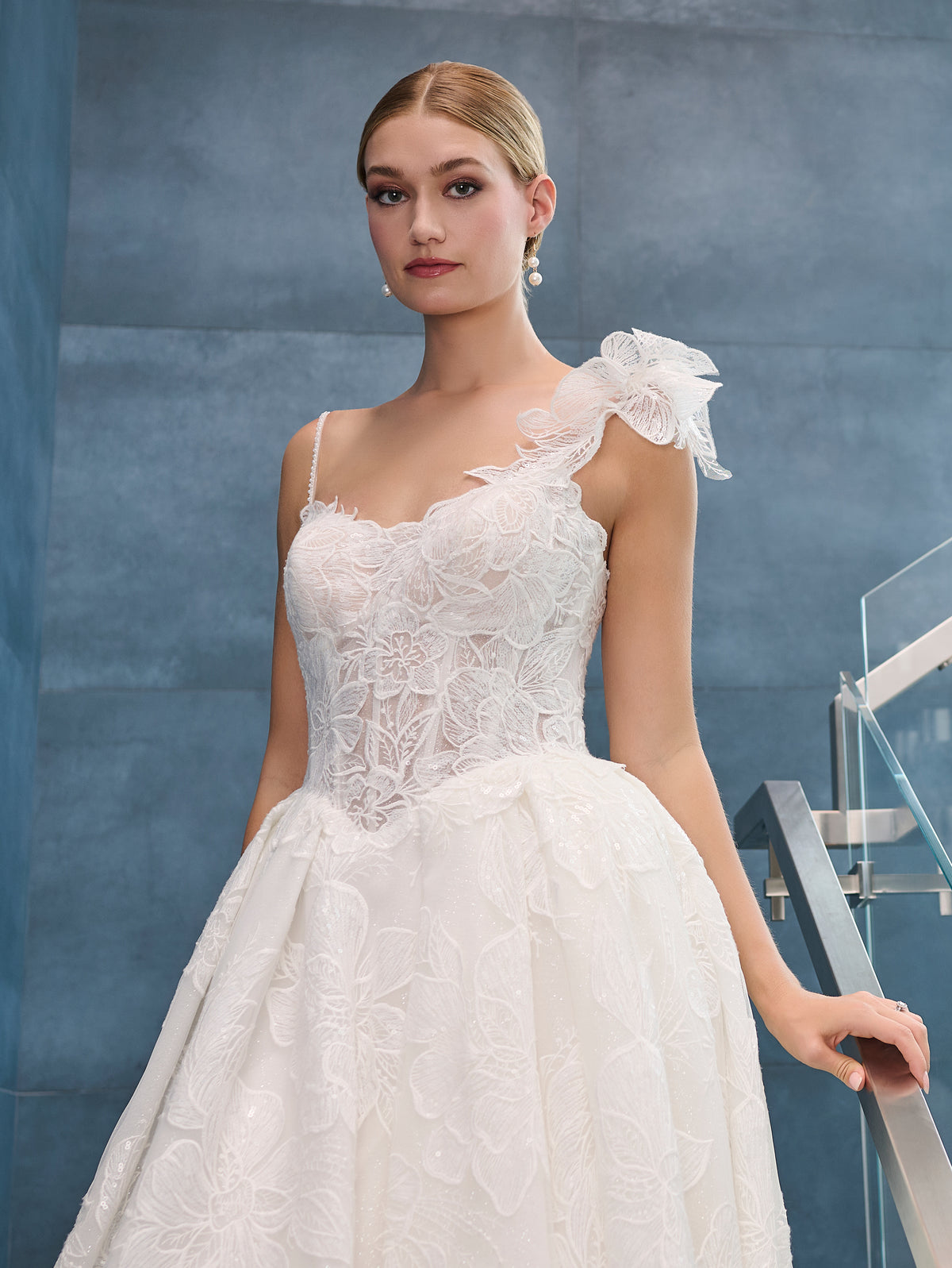 A woman modeling a white one-shoulder wedding dress with floral lace embroidery, a sweetheart neckline, and a ruffled bow detail on the shoulder, set against a textured blue-gray wall. / This is a stunning ball gown made out of floral all over lace with glitter mesh underlayer. Semi-sheer bodice with a scooped neckline, beaded spaghetti straps and beautiful lace feature on one shoulder.
