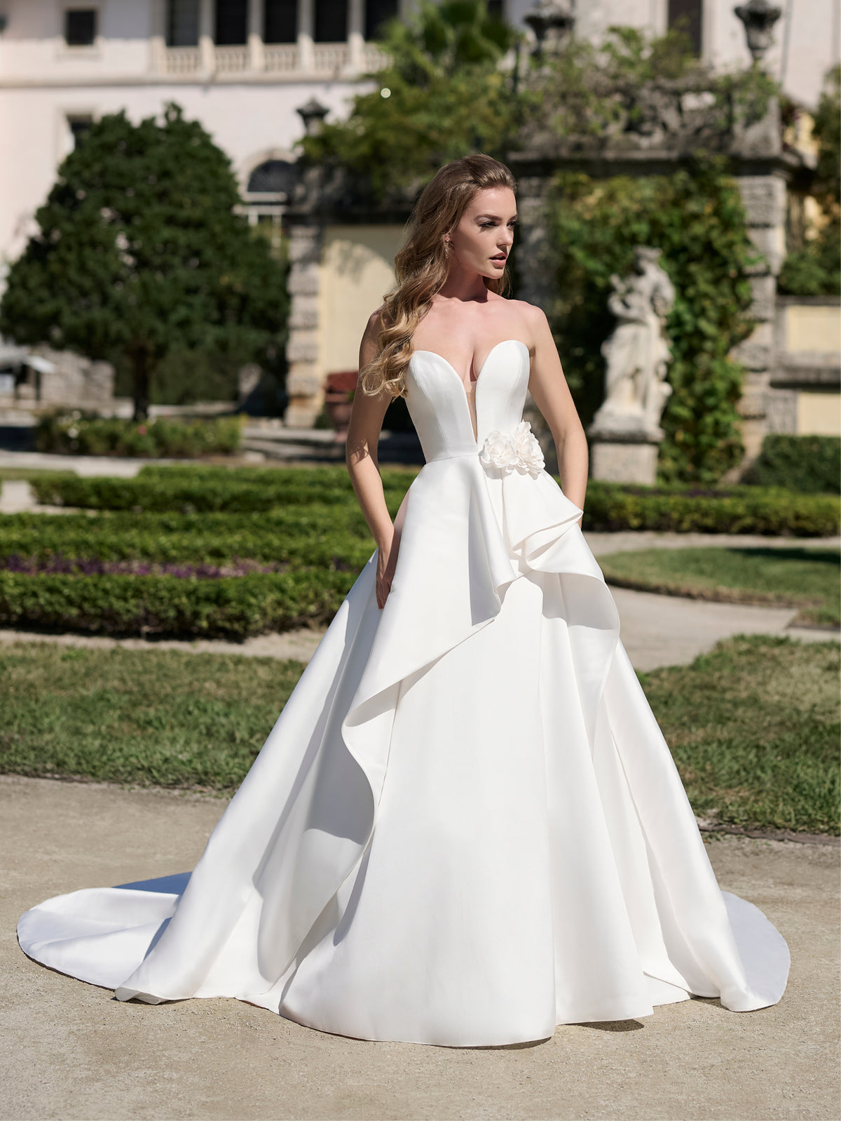 Woman in a white wedding dress standing in an elegant garden setting / Strapless satin ballgown with 3D flowers and sculpted cascade on the skirt.