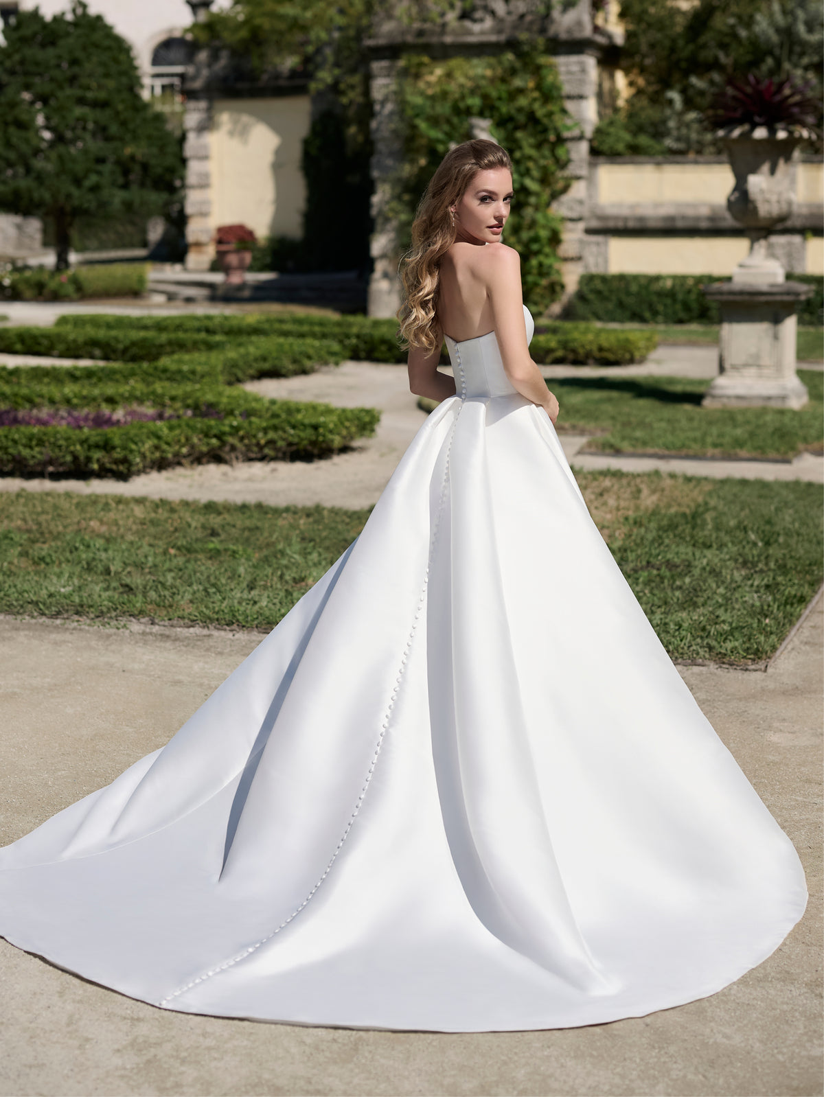 Woman in a white wedding dress standing outdoors in a garden setting / Strapless satin ballgown with 3D flowers and sculpted cascade on the skirt.