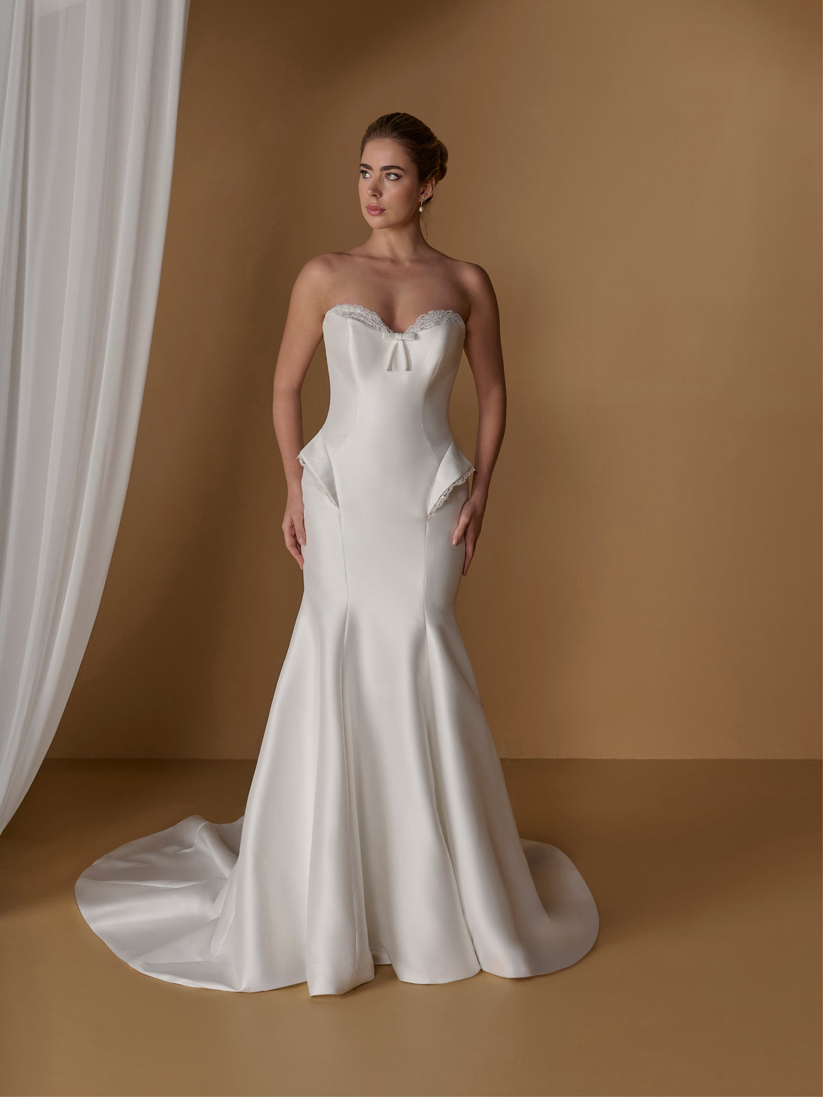 A woman modeling a strapless white mermaid-style wedding gown with a sweetheart neckline, fitted bodice, and full skirt with a train, posed against a warm tan studio backdrop with draped white fabric at the side. / Stretch mikado slim gown with lace and peplum detailing and lace up back.
