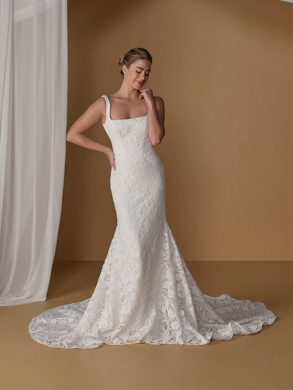 A woman modeling a fitted white lace wedding gown with thin straps, a square neckline, and a long train in a warm tan studio setting with a white curtain on the left. / All over lace gown with square neckline.