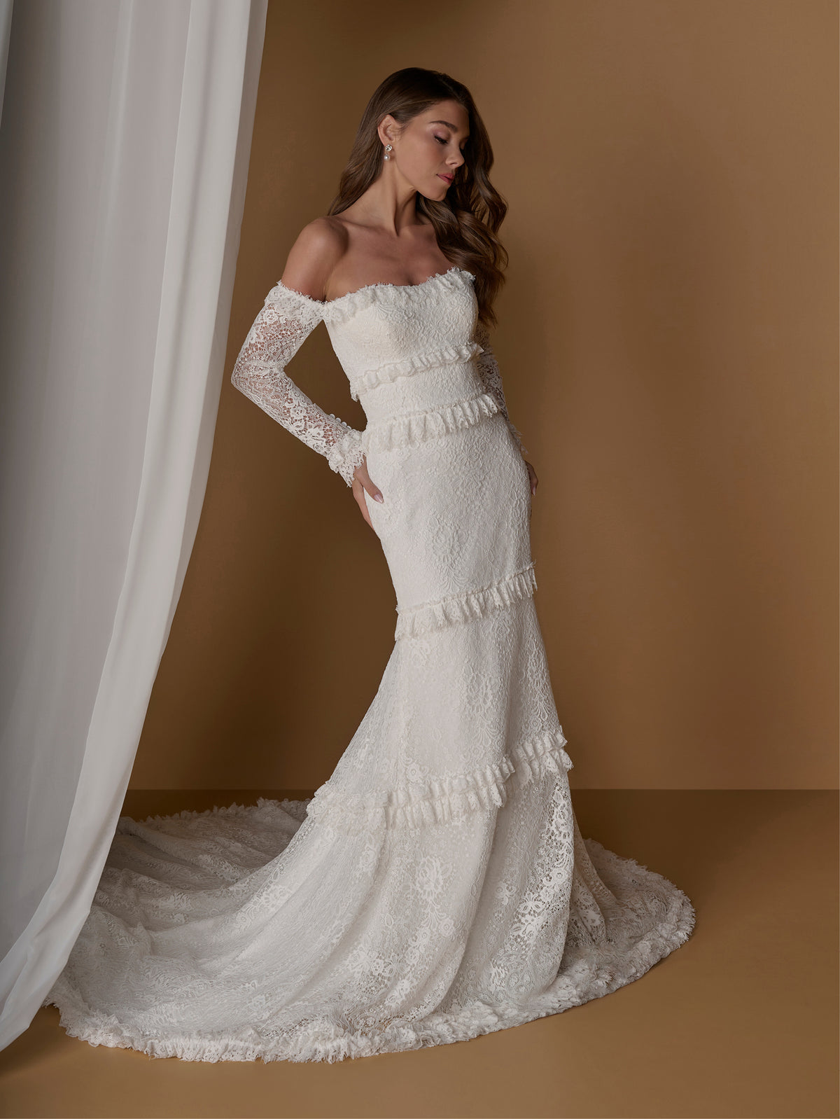 An off-the-shoulder lace wedding gown with long sheer sleeves and a flared mermaid silhouette with ruffle trim, shown on a model against a warm brown studio backdrop with a white curtain. / Slim, long sleeve lace gown with lace ruffle detail.