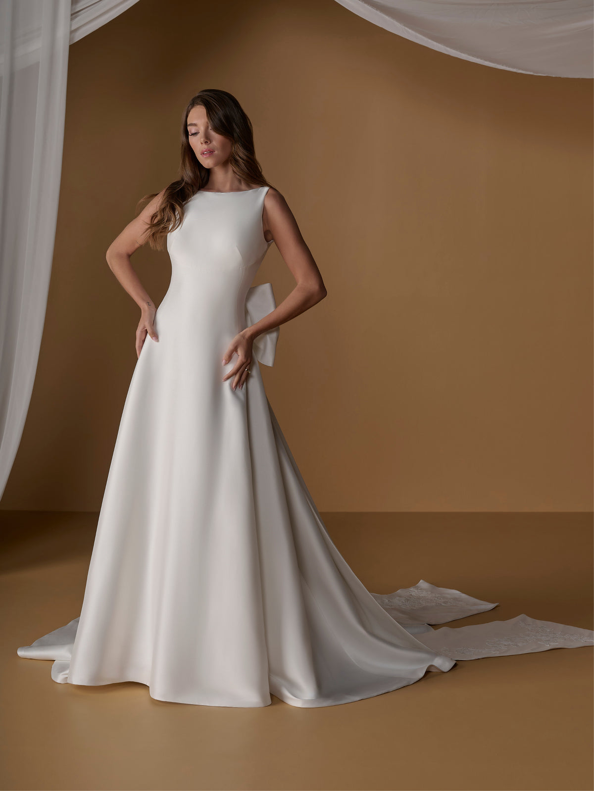A woman models a sleeveless white wedding gown with a high neckline, fitted bodice, and full satin-like skirt with a long train, posed against a warm tan studio backdrop with draped white fabric. / A-line ballgown with modern dart detailing, illusion back and detachable bow.