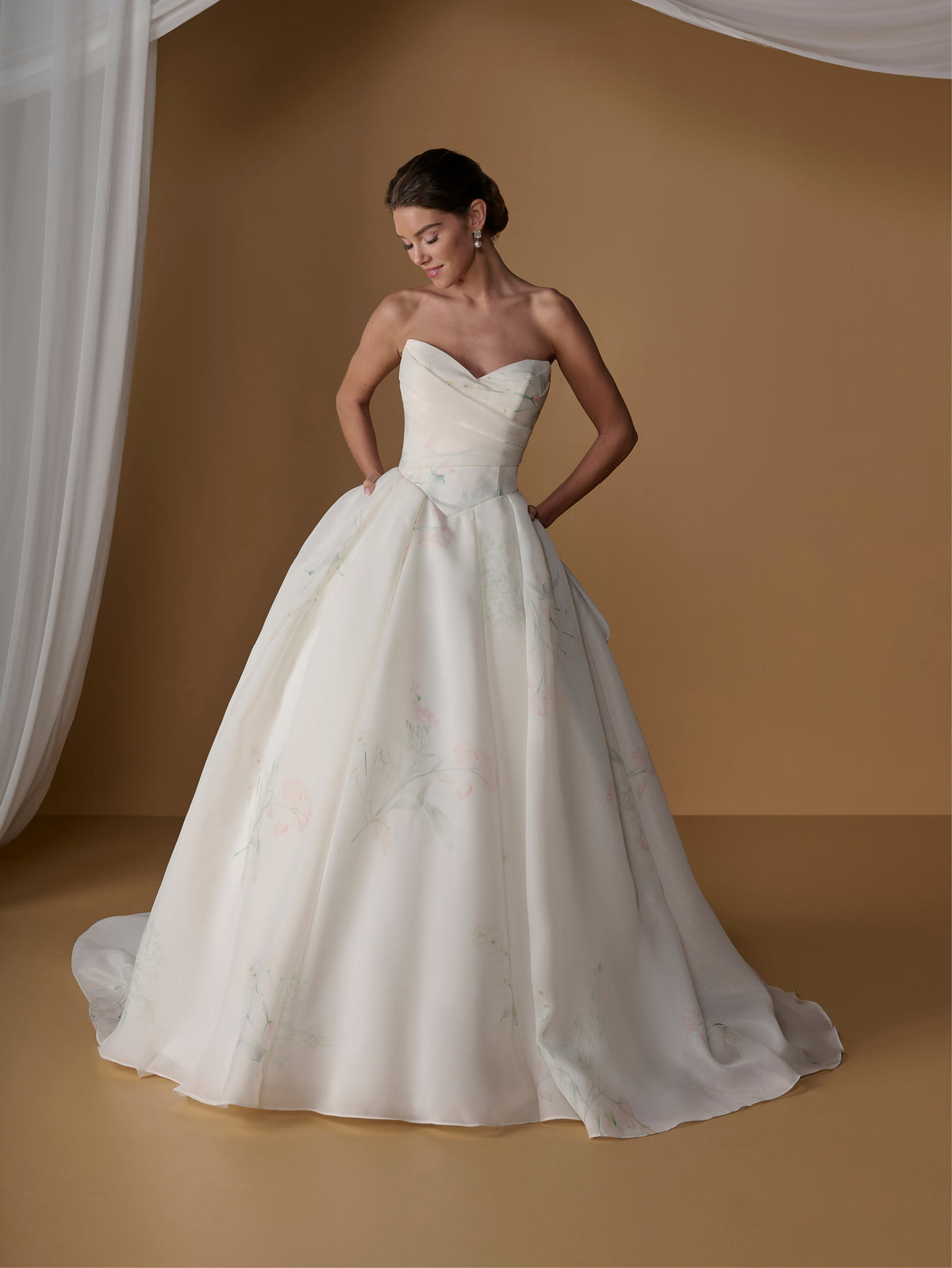 A woman modeling a strapless sweetheart ball gown wedding dress with a full skirt and subtle floral embroidery, posed against a warm tan studio backdrop with draped white fabric on the left. / Satin faced organza printed ballgown with dramatic skirt and bow jacket.