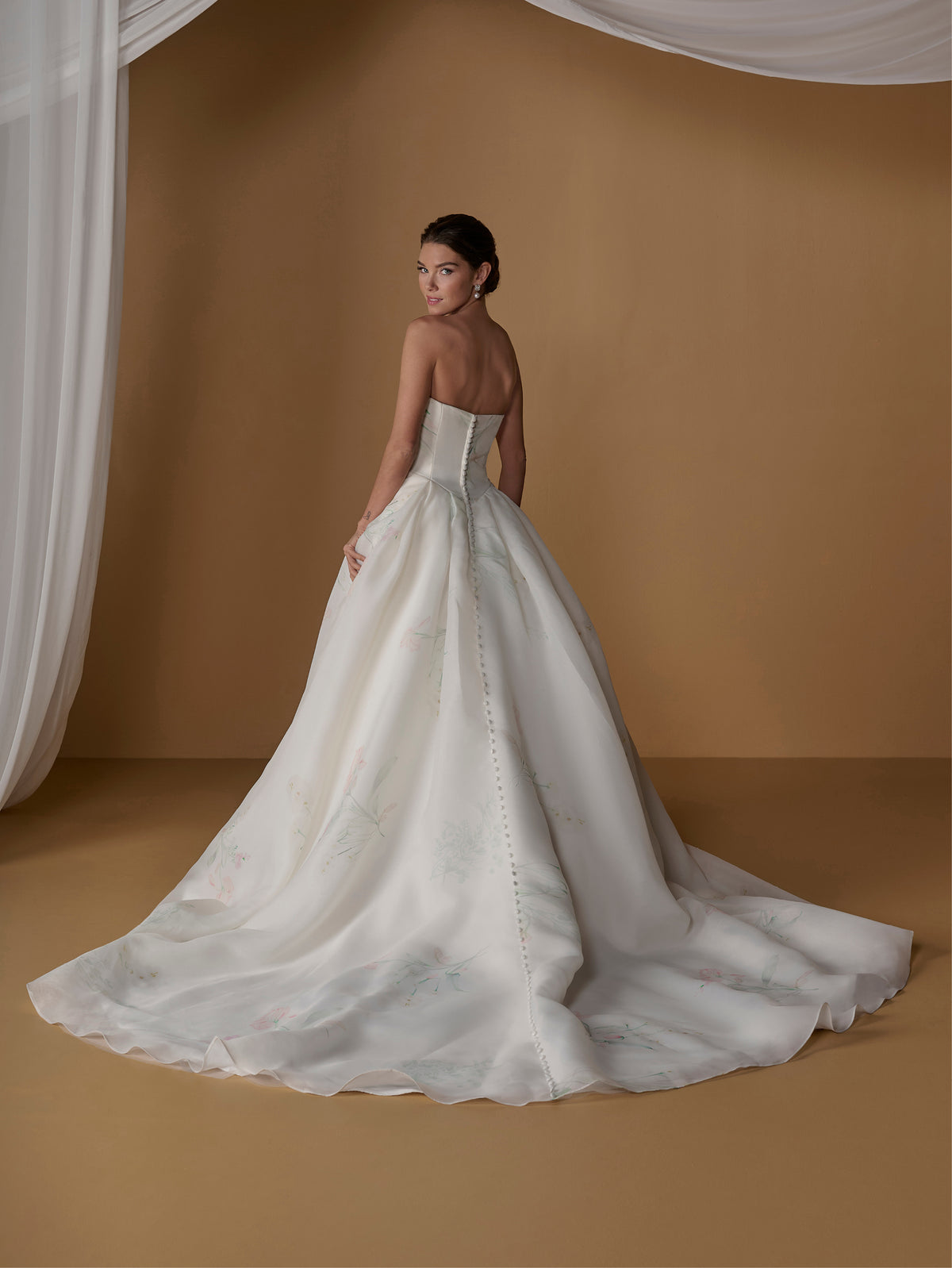 A woman wearing a strapless white ball gown wedding dress with a structured corset-style bodice and a full flowing skirt, photographed in a warm tan studio setting with draped white fabric at the top. / Satin faced organza printed ballgown with dramatic skirt and bow jacket.
